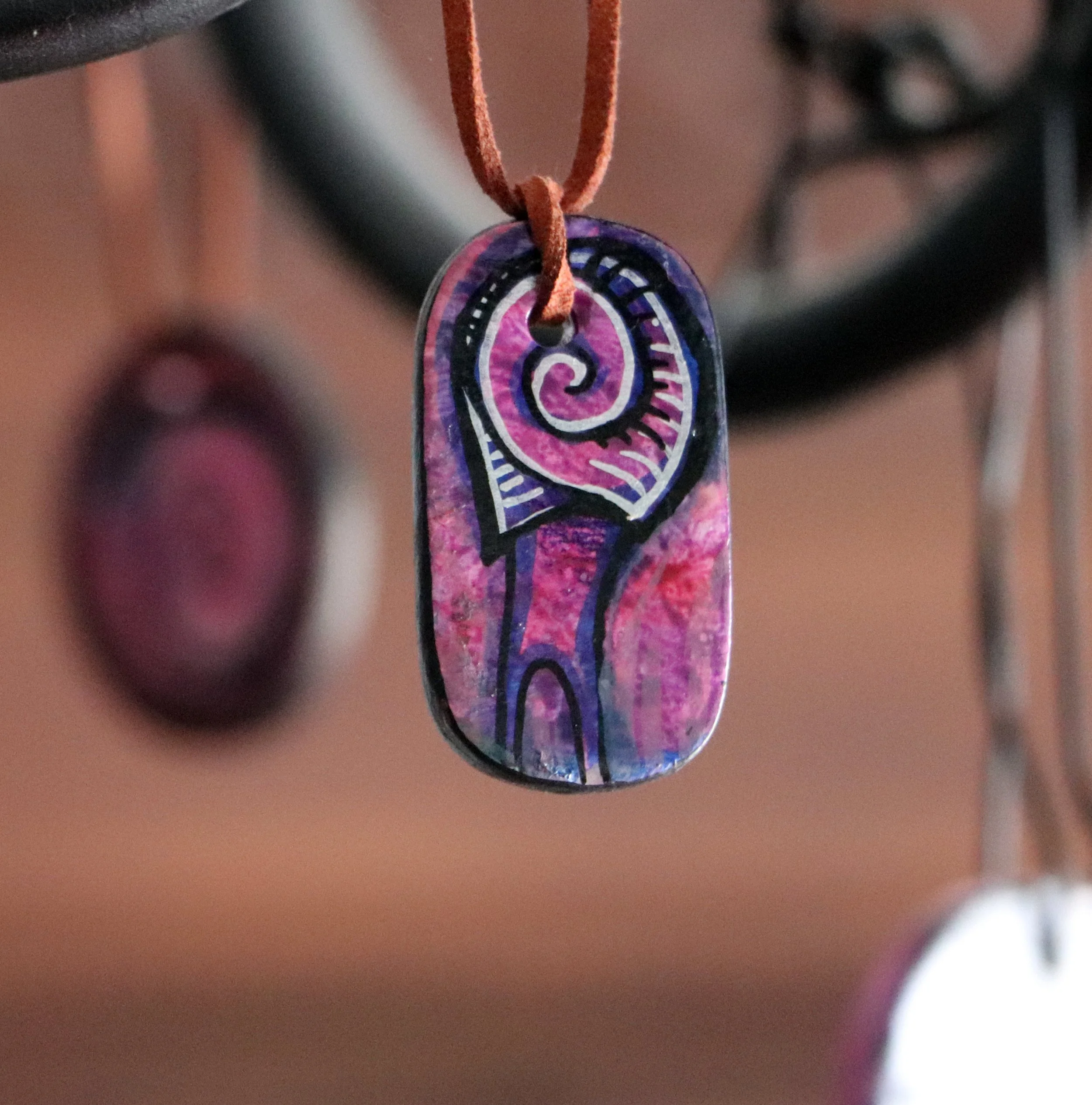 Hand-painted ceramic pendant with a purple and pink spiral design hanging from a leather cord, blurred background.