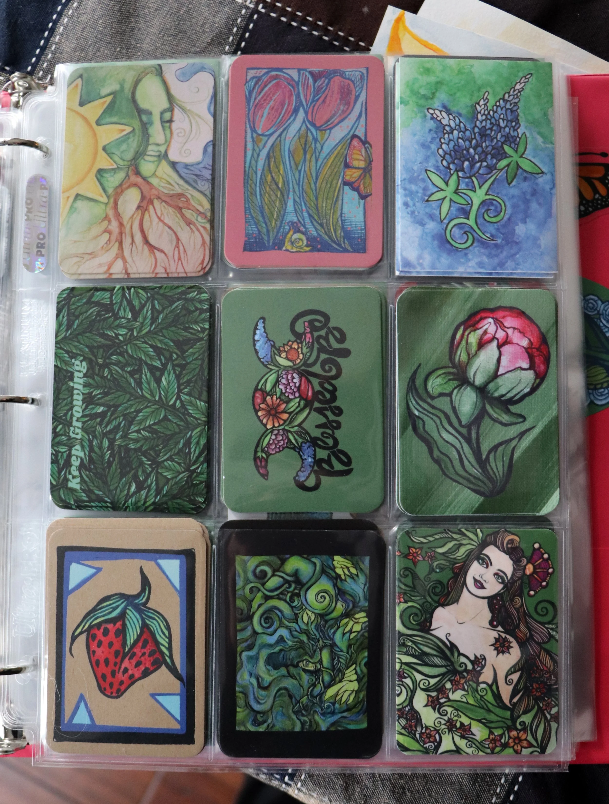 Collection of nine illustrated cards in a plastic album, featuring floral, nature, and artistic designs, including a woman with plant-like features, flowers, and abstract patterns.