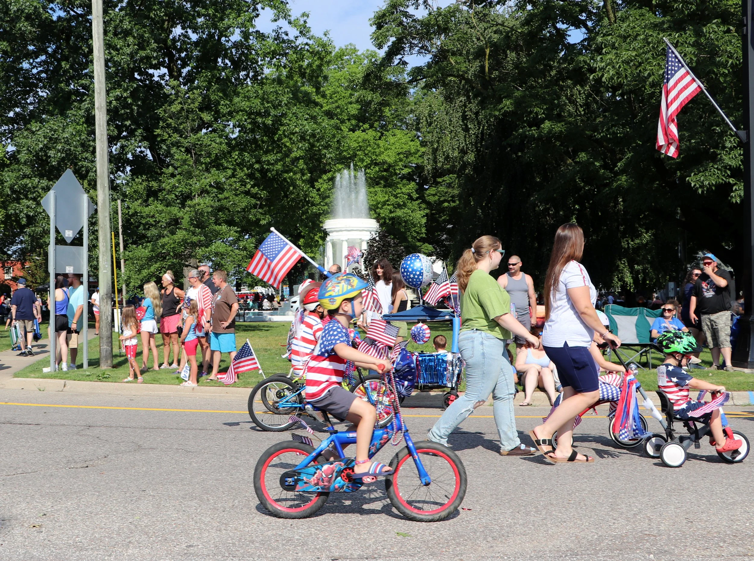 A crowd celebrating outdoors with American flags, some children riding bikes and wearing patriotic clothing, in front of green trees and a fountain.