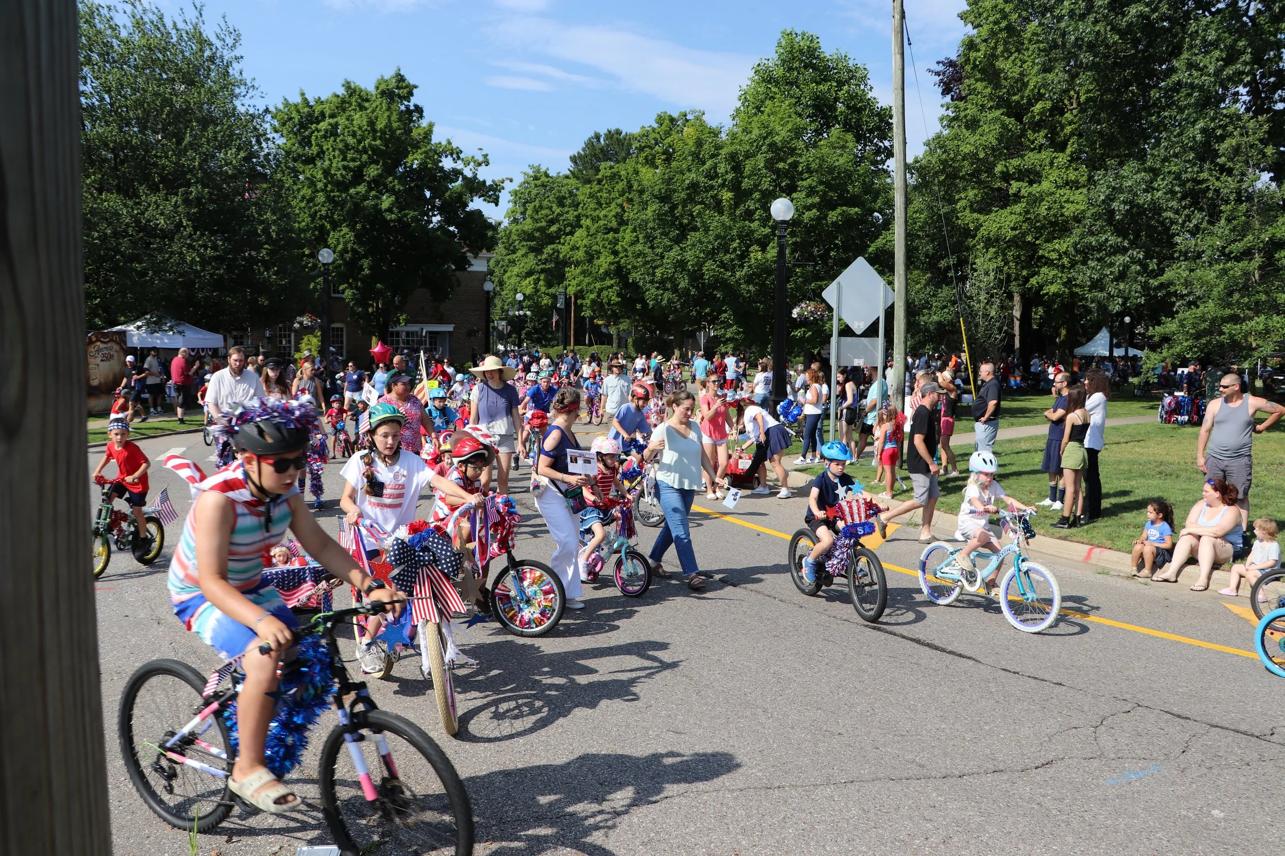 A parade with children riding decorated bicycles and people watching on the sidewalk, patriotic decorations and American flags in a park setting on a sunny day.