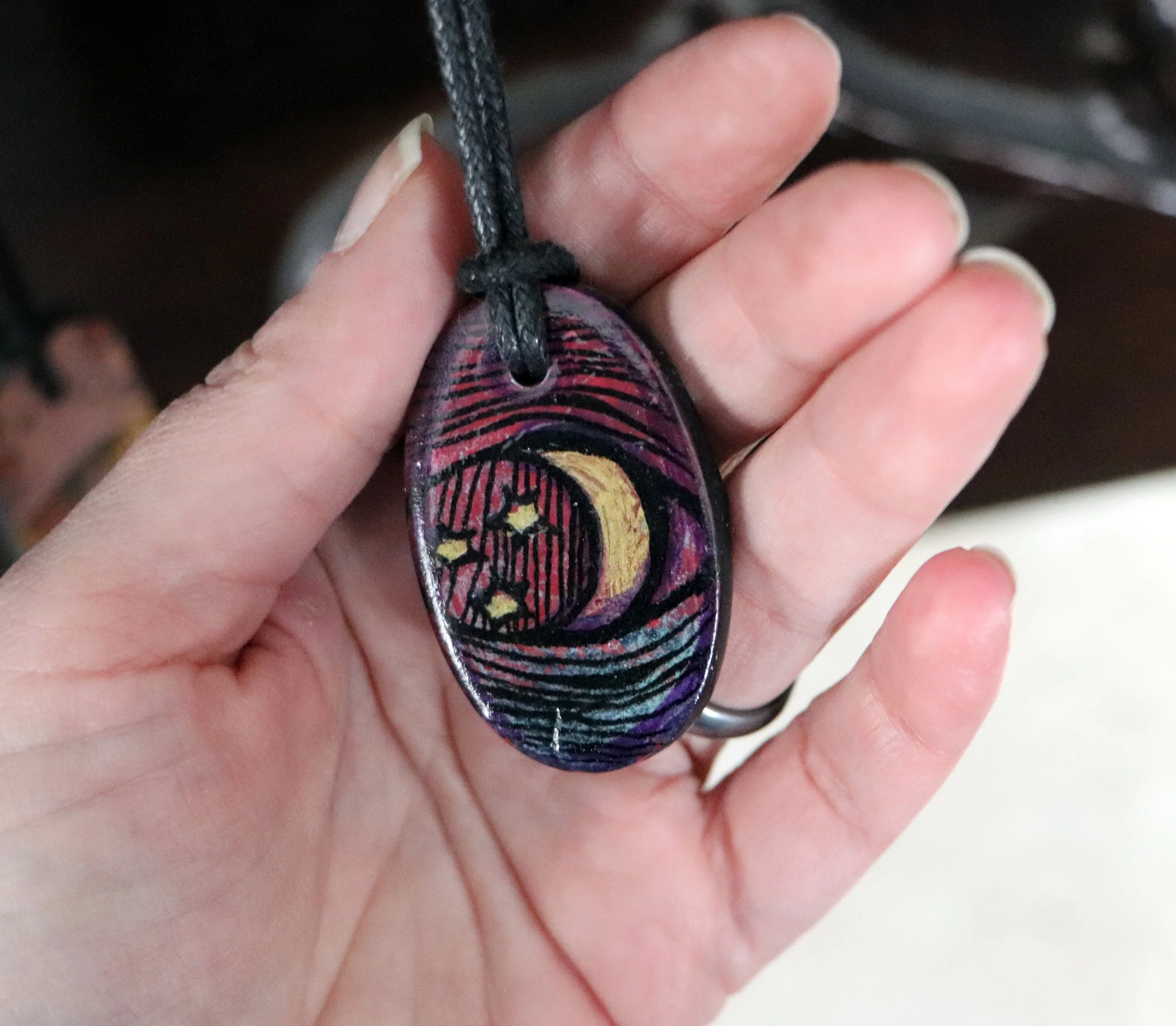Hand holding a pendant with a painted night sky featuring a crescent moon and stars on a black cord