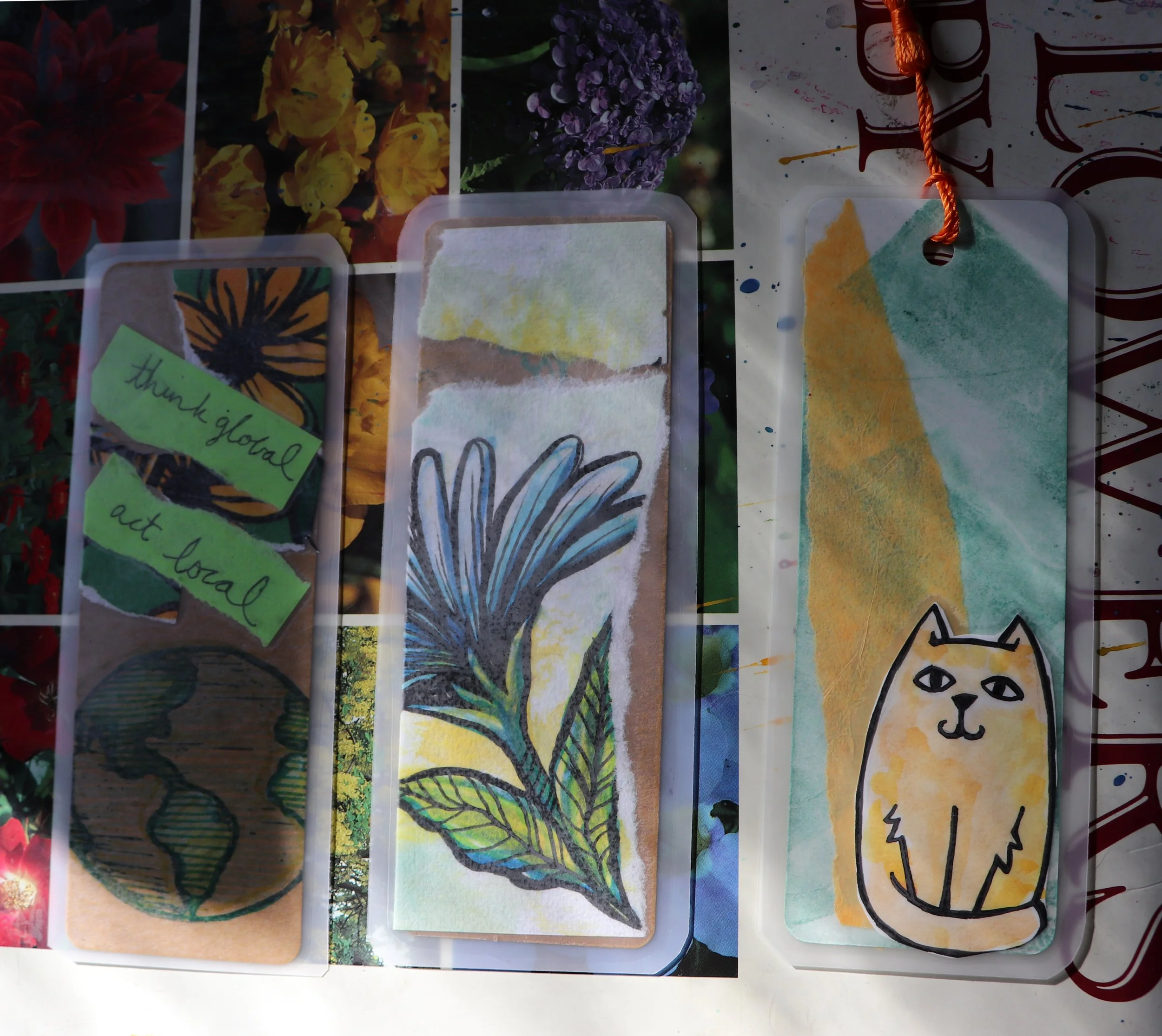 Three bookmarks with nature-themed illustrations and messages, including a globe with text 'think global act local,' a flower, and a yellow background with a cat drawing, hanging on an orange string.