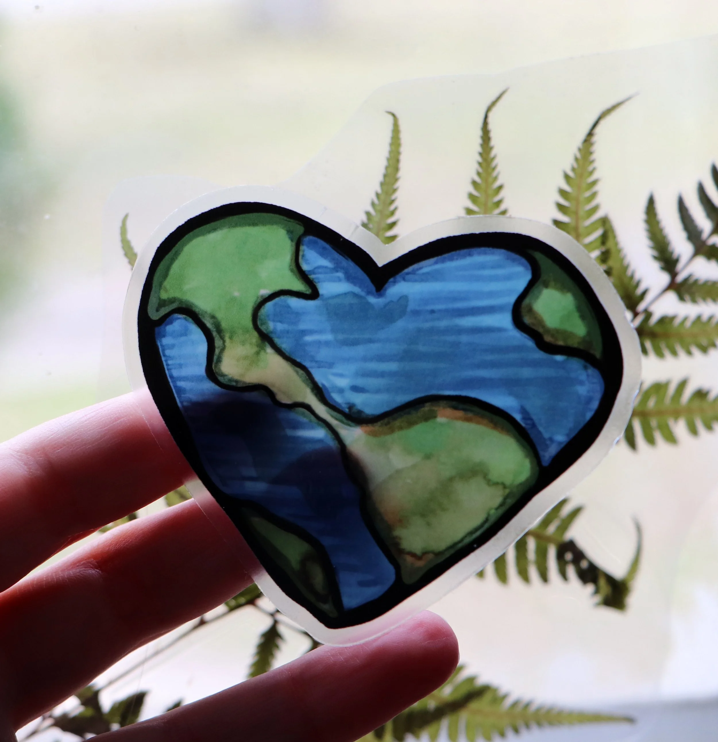 Close-up of a colorful sticker featuring a stylized Earth with green continents and blue oceans, held by a person's fingers, with green fern leaves in the background.