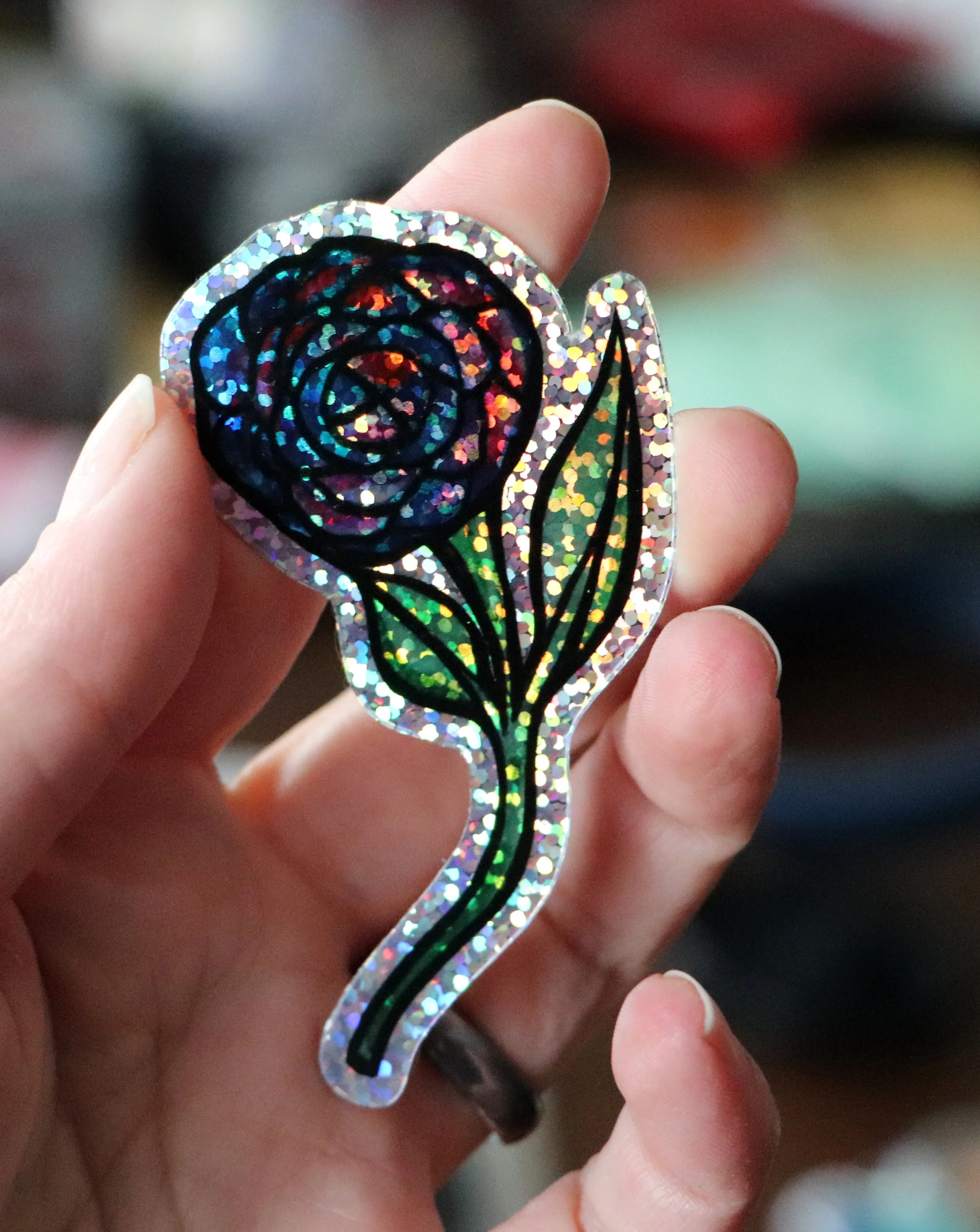 A hand holding a decorative hair clip with a glittery, holographic surface and a black, floral design in the center.