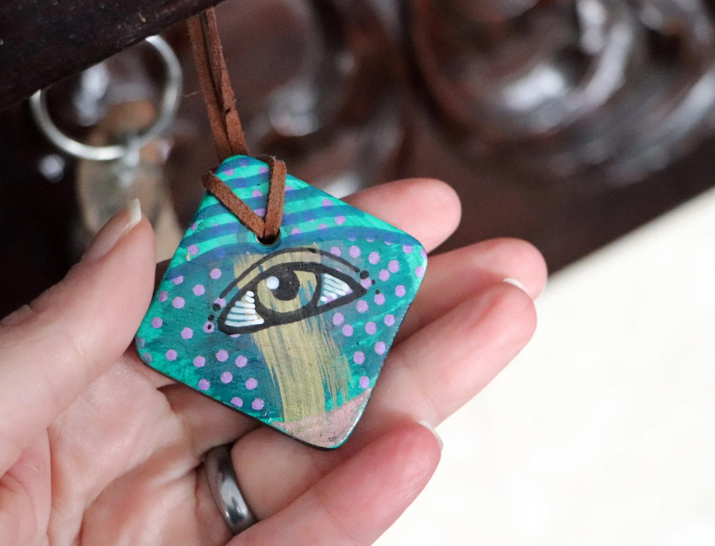 A hand holding a painted square pendant featuring an eye drawing with a gold iris, white details, and a black outline, on a blue background with purple dots, attached to a brown leather cord, near a dark wooden surface with hooks and keys.