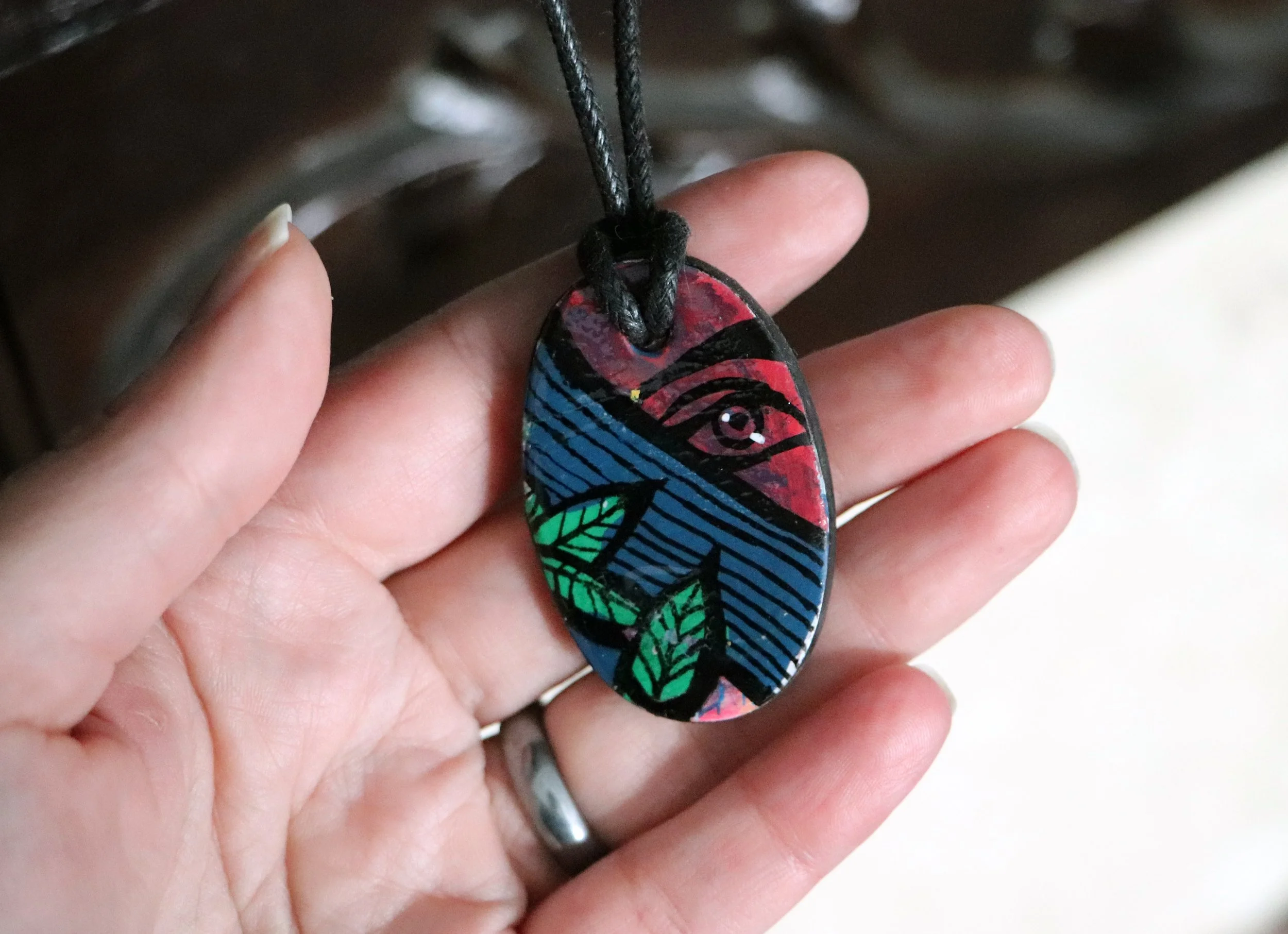 A hand holding an oval-shaped pendant with a colorful, abstract design of an eye, leaves, and lines, attached to a black cord.