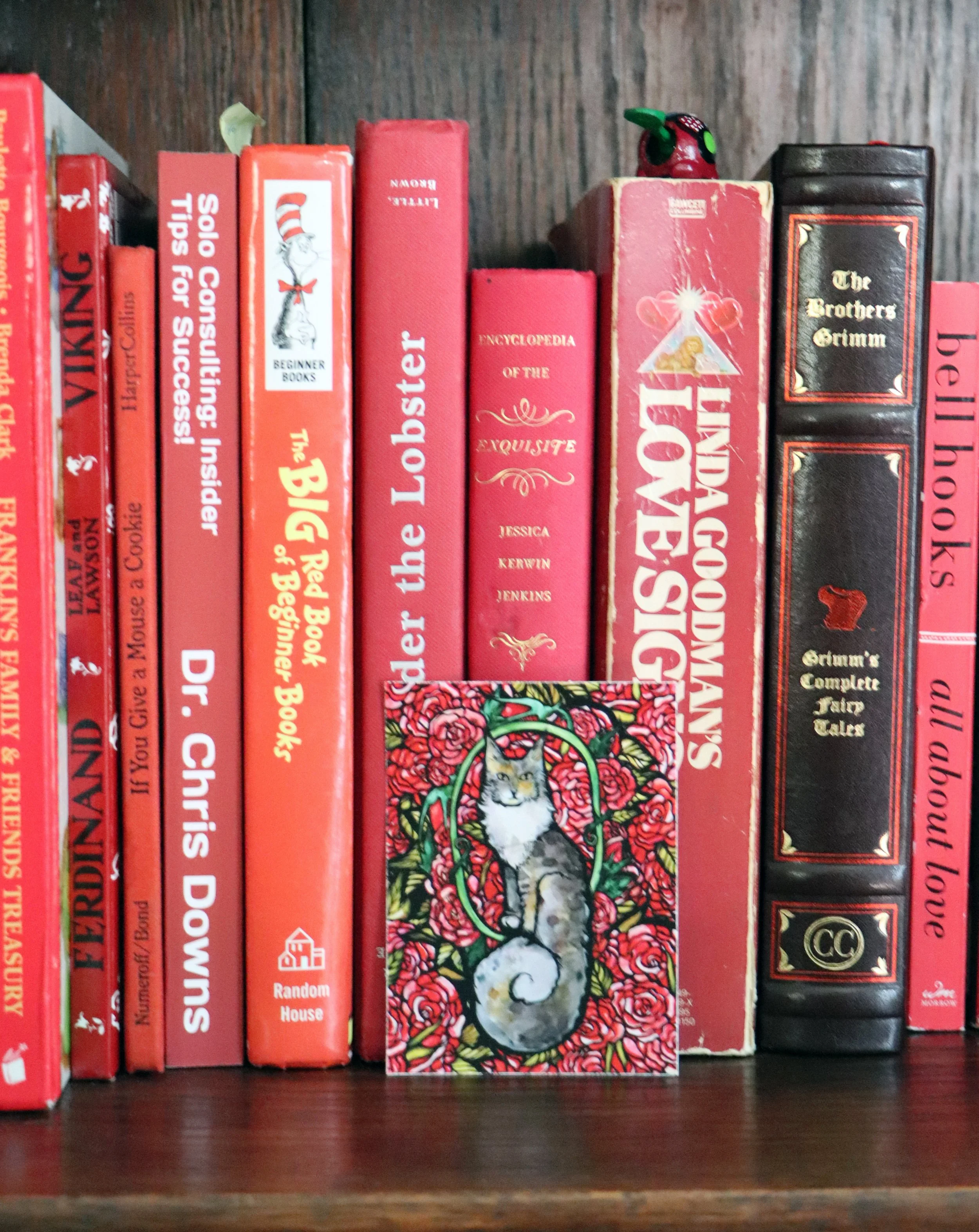 A row of books on a wooden shelf with a decorative card featuring a cat surrounded by red roses in front of the books.