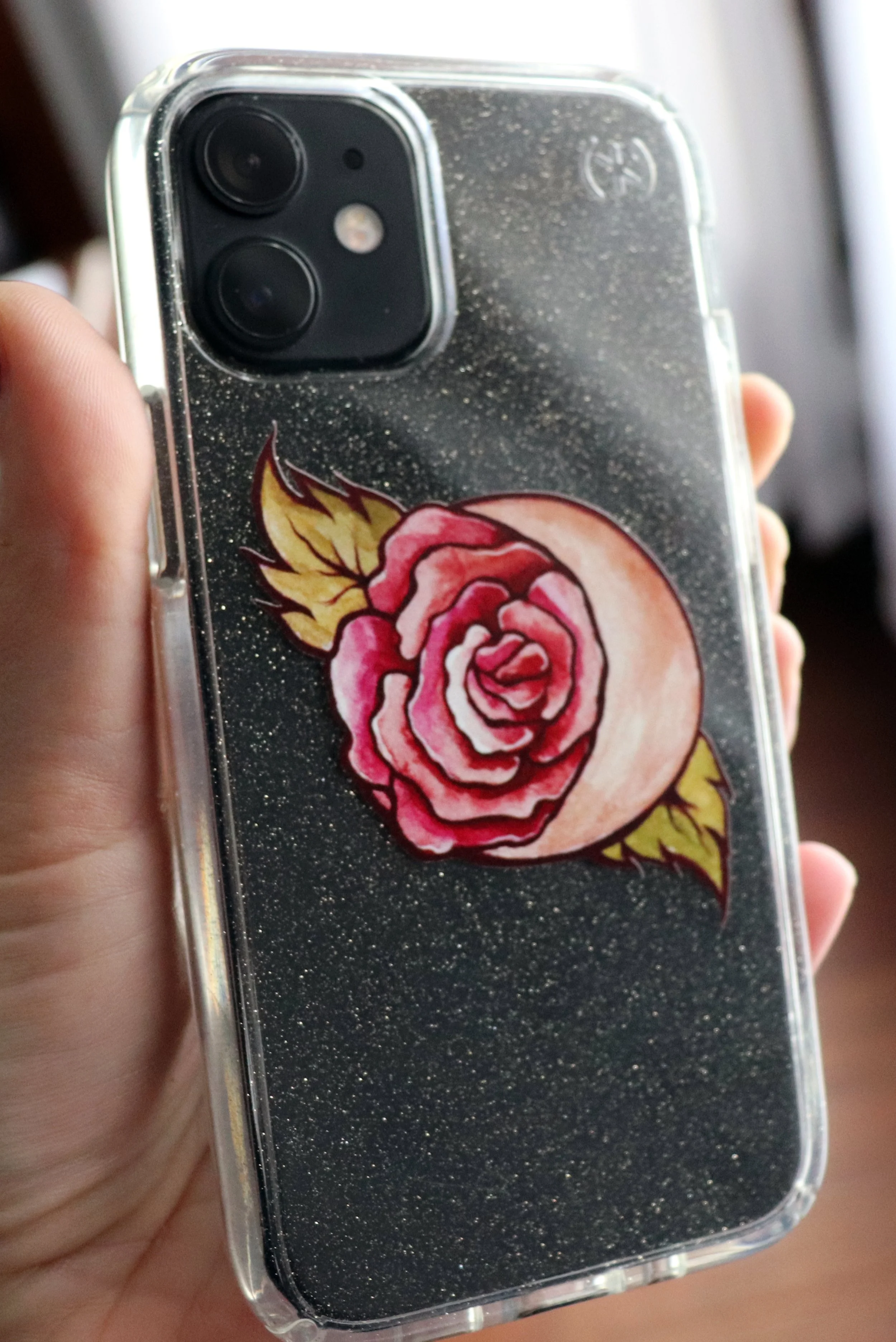 Close-up of a person holding a smartphone with a black, glittery case that has a pink and yellow rose design.