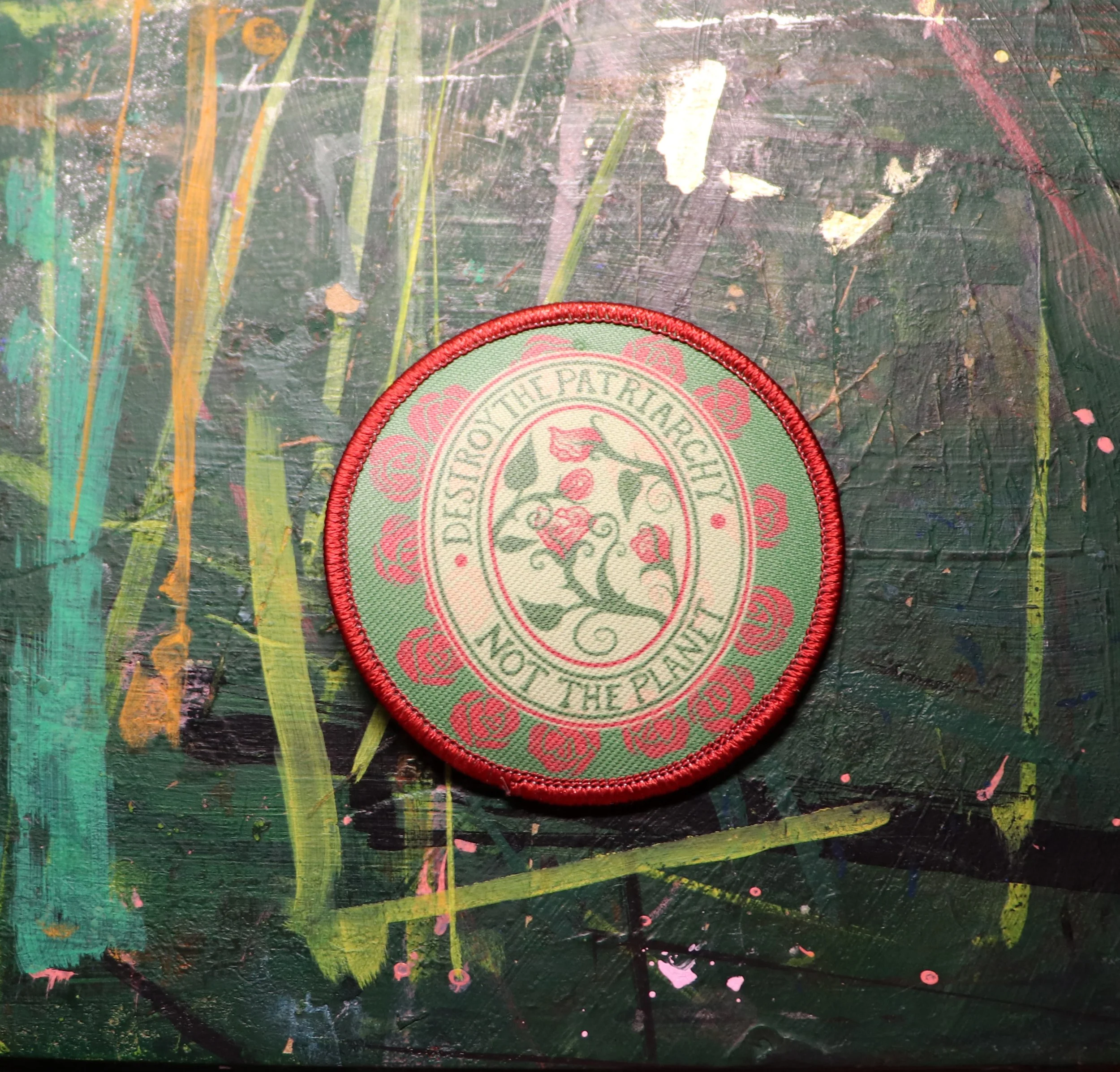 A circular embroidered patch with a green background, a floral design in the center, pink roses, and a red border. The patch has the words "DESIGN THE PATH, NOT THE PLANET" encircling the floral design.