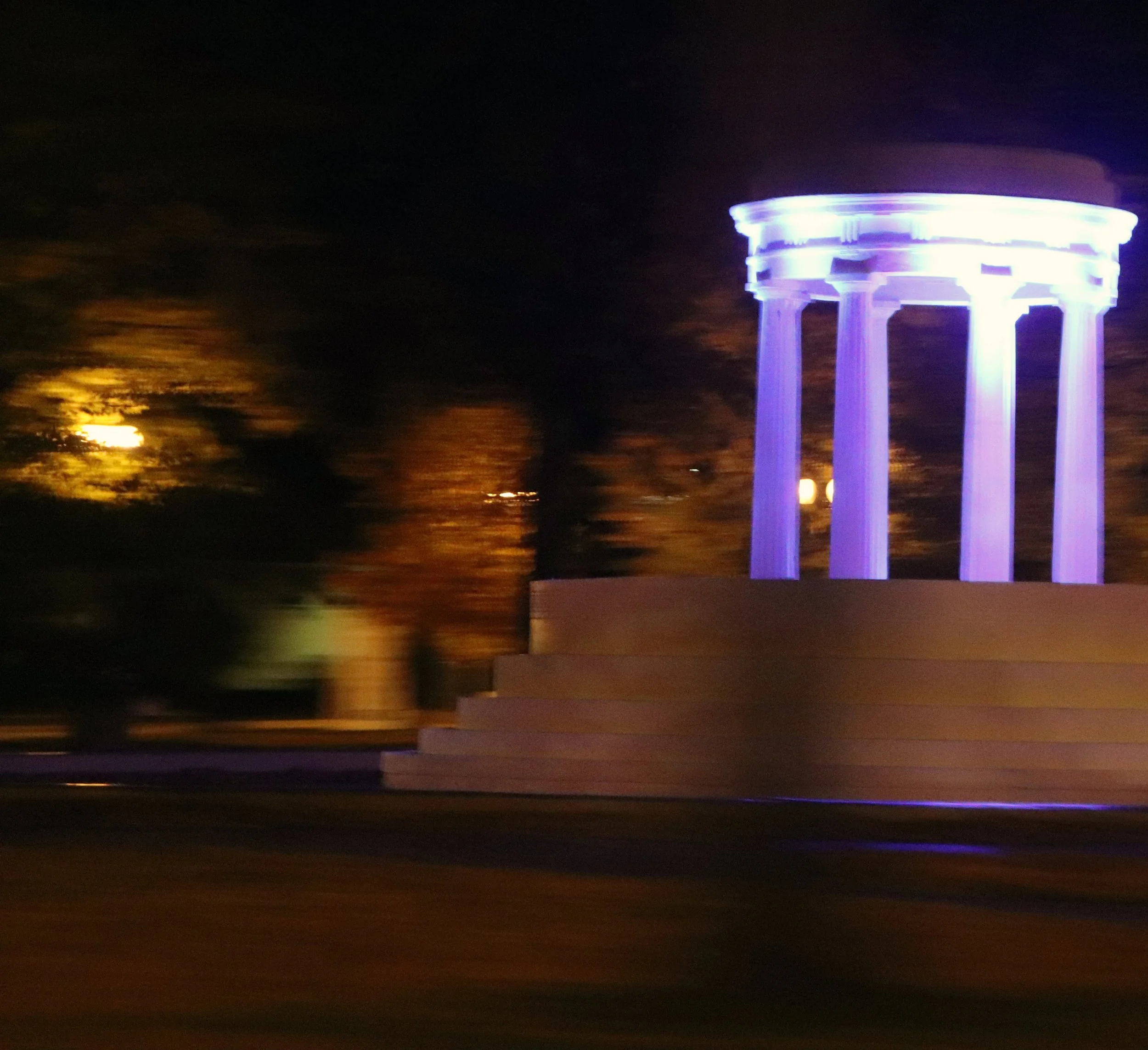 Nighttime view of the Virginia State Capitol building illuminated with purple and blue lights, with trees and blurred moving lights in the background.