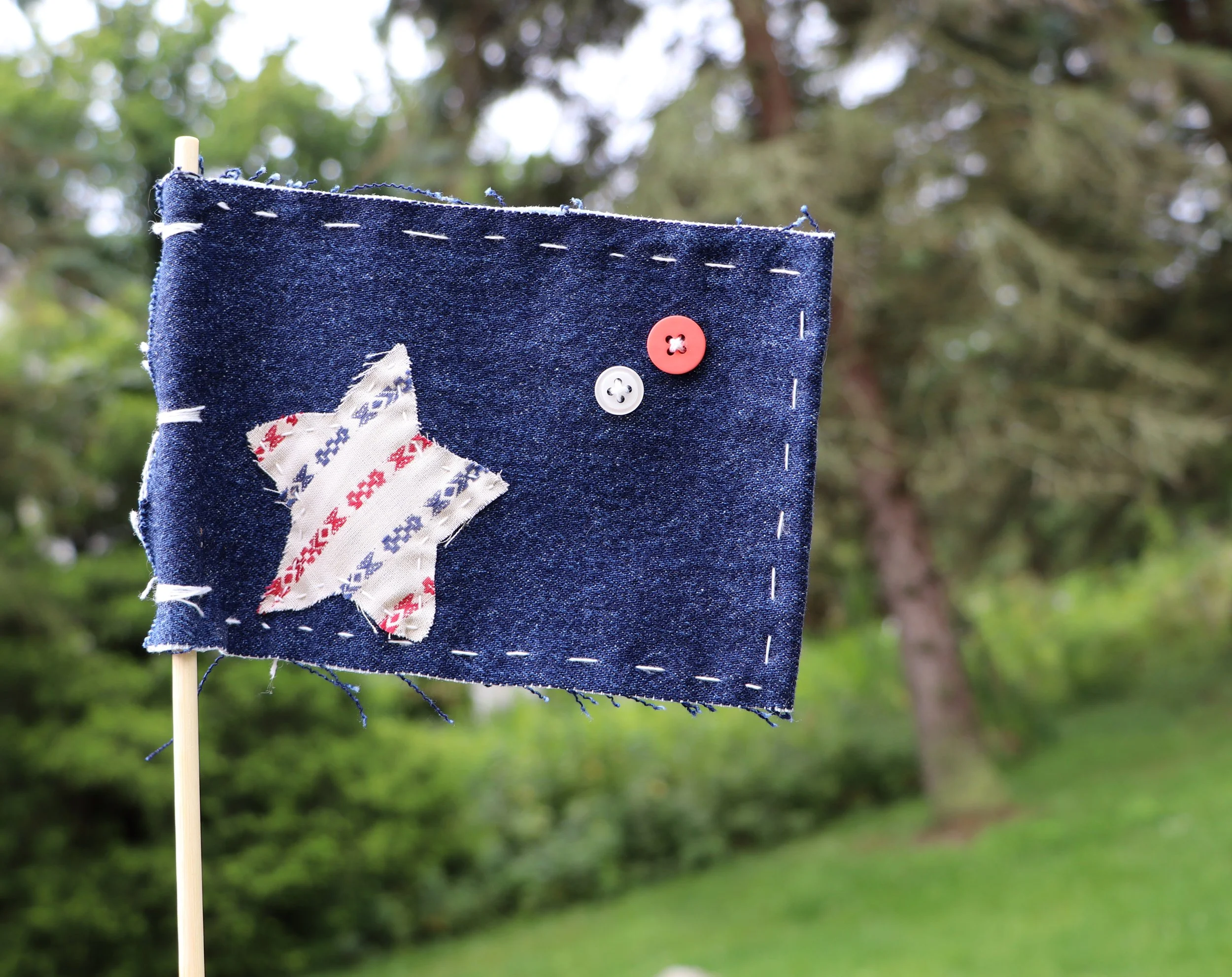 A piece of denim fabric with frayed edges and stitched seams, attached to a wooden stick with white and red buttons, set outdoors with blurred trees and greenery in the background.