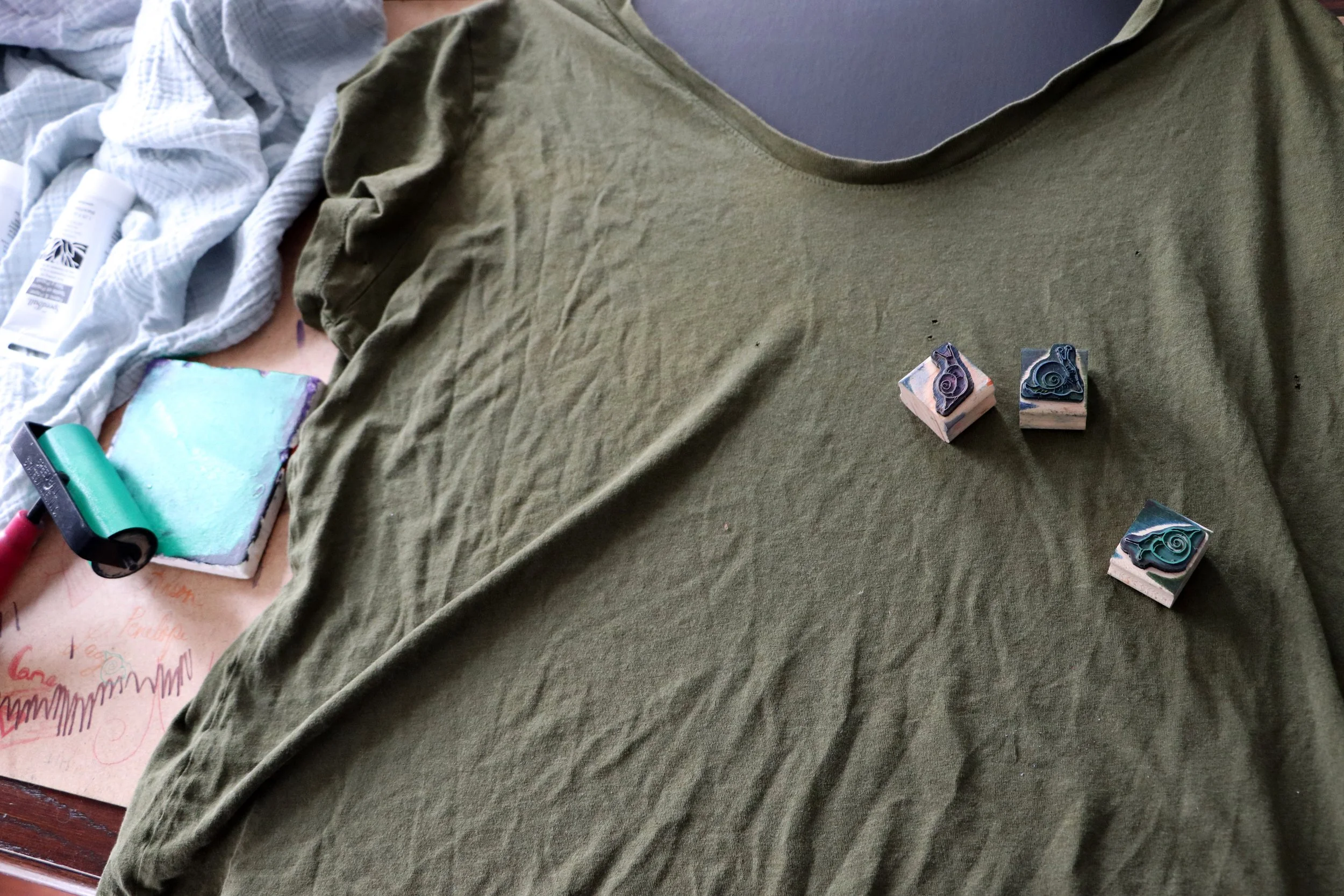 Green T-shirt with three multicolored snail stamps on it. A soap carving tool and a soap block with carved soap in the background.