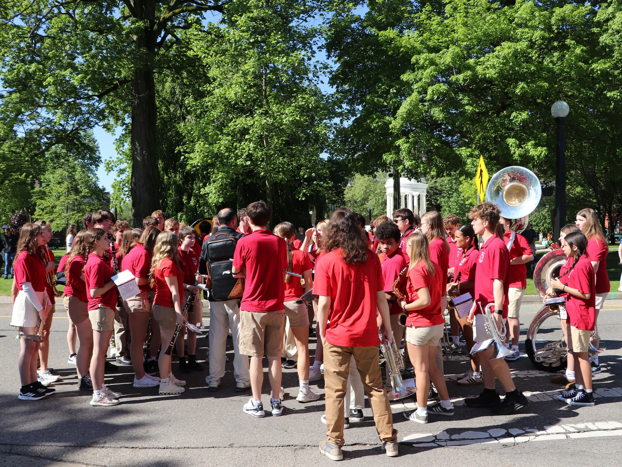A marching band dressed in red shirts prepares to perform outdoors on a sunny day, with lush green trees and a white pavilion in the background.
