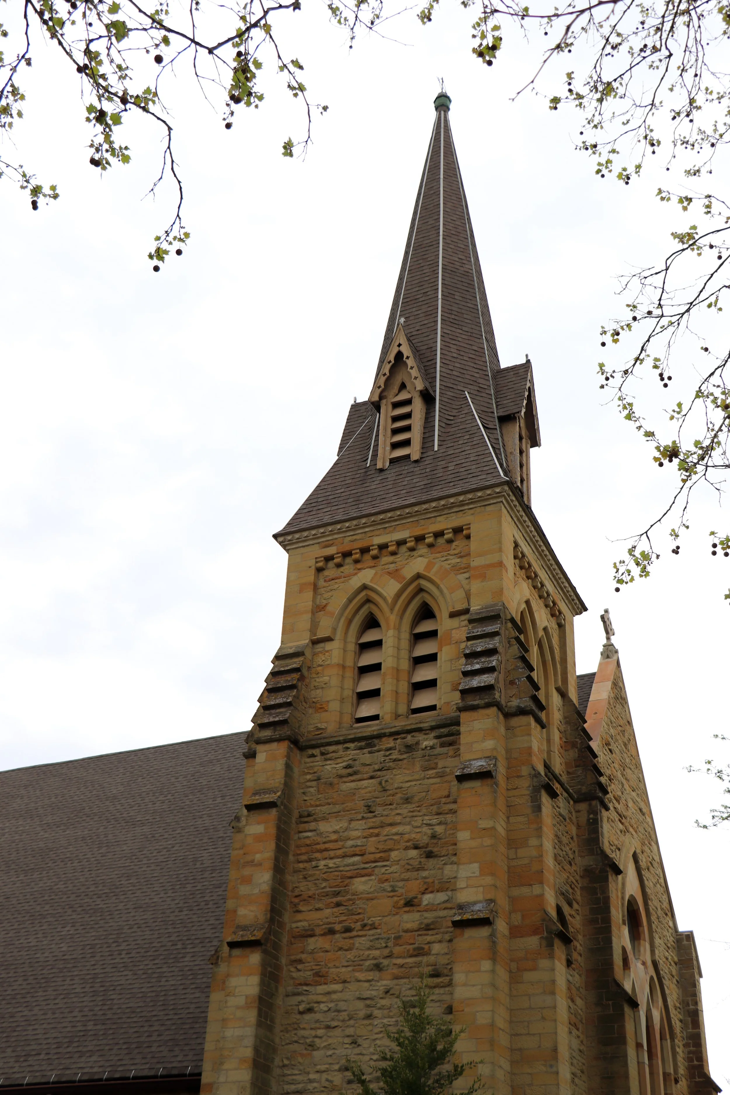 View of a tall church steeple with a pointed spire, set against an overcast sky. The church's architecture features brick and stonework with arched windows and decorative elements, partially framed by tree branches with leaves.