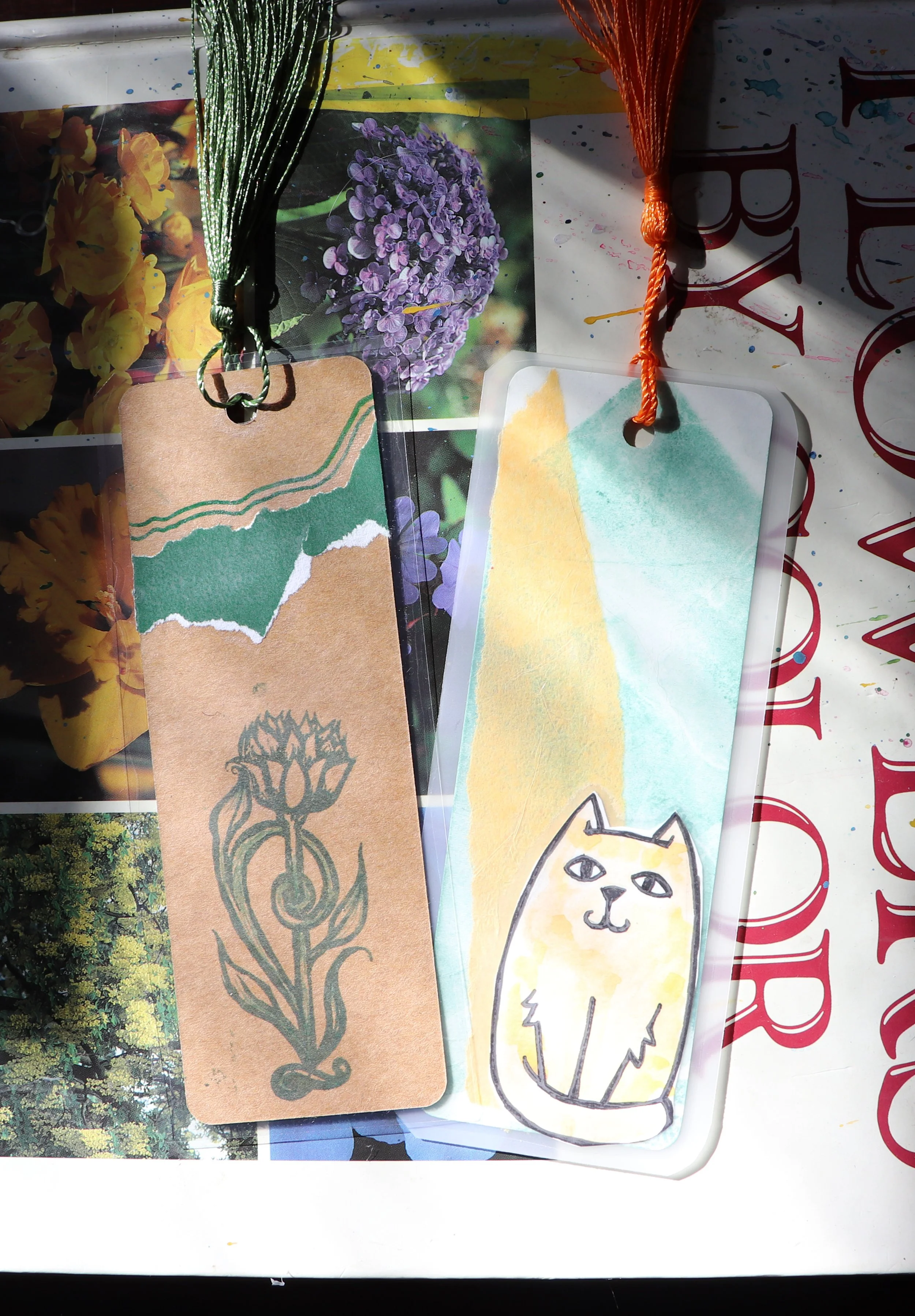 Two handmade bookmarks with string ties on a collage of photographs and materials. One bookmark has green and beige torn paper with a black and gray illustrated flower, the other has a pastel watercolor background with a simple drawing of a cat.