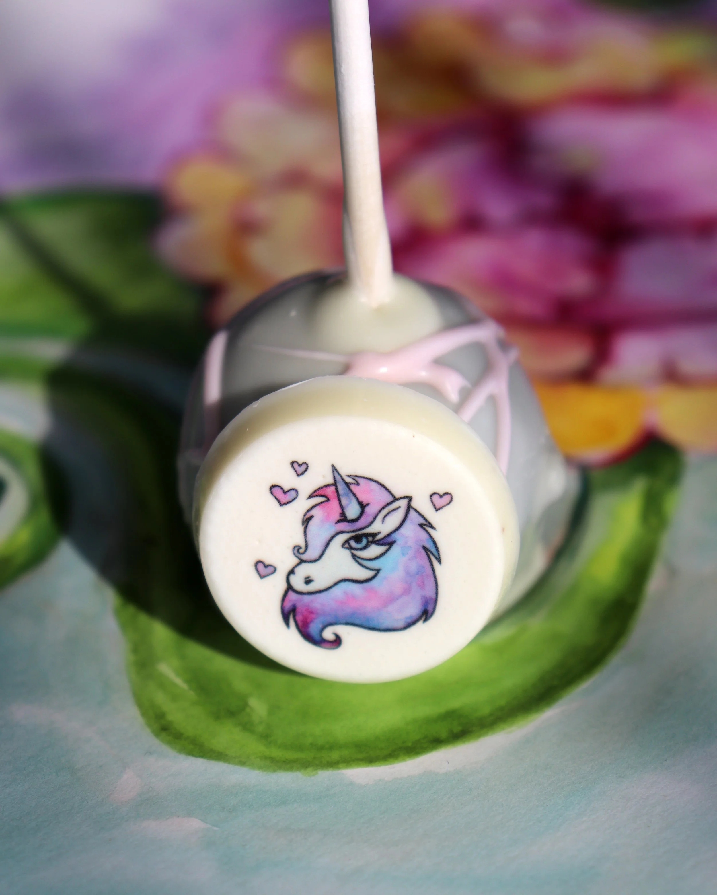 Close-up of a watermelon-shaped object with a small circular tag featuring a cartoon unicorn with a rainbow-colored mane and a horn, surrounded by pink hearts.