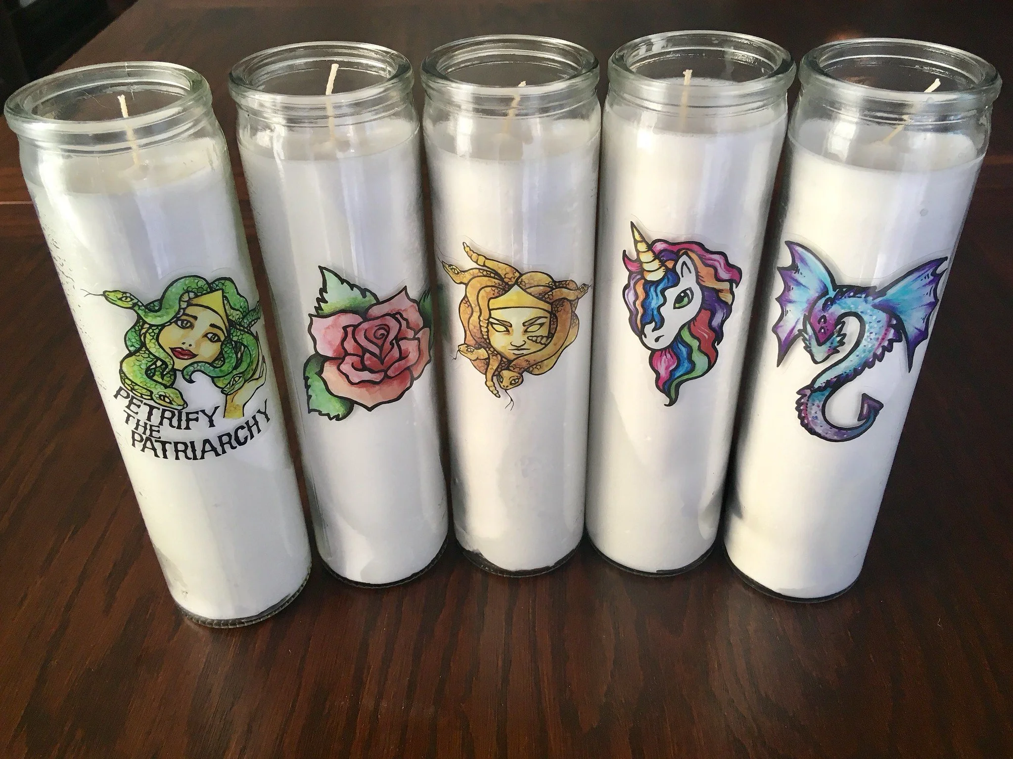 Five candles in glass jars with colorful sticker designs featuring fantasy characters, placed on a wooden surface.