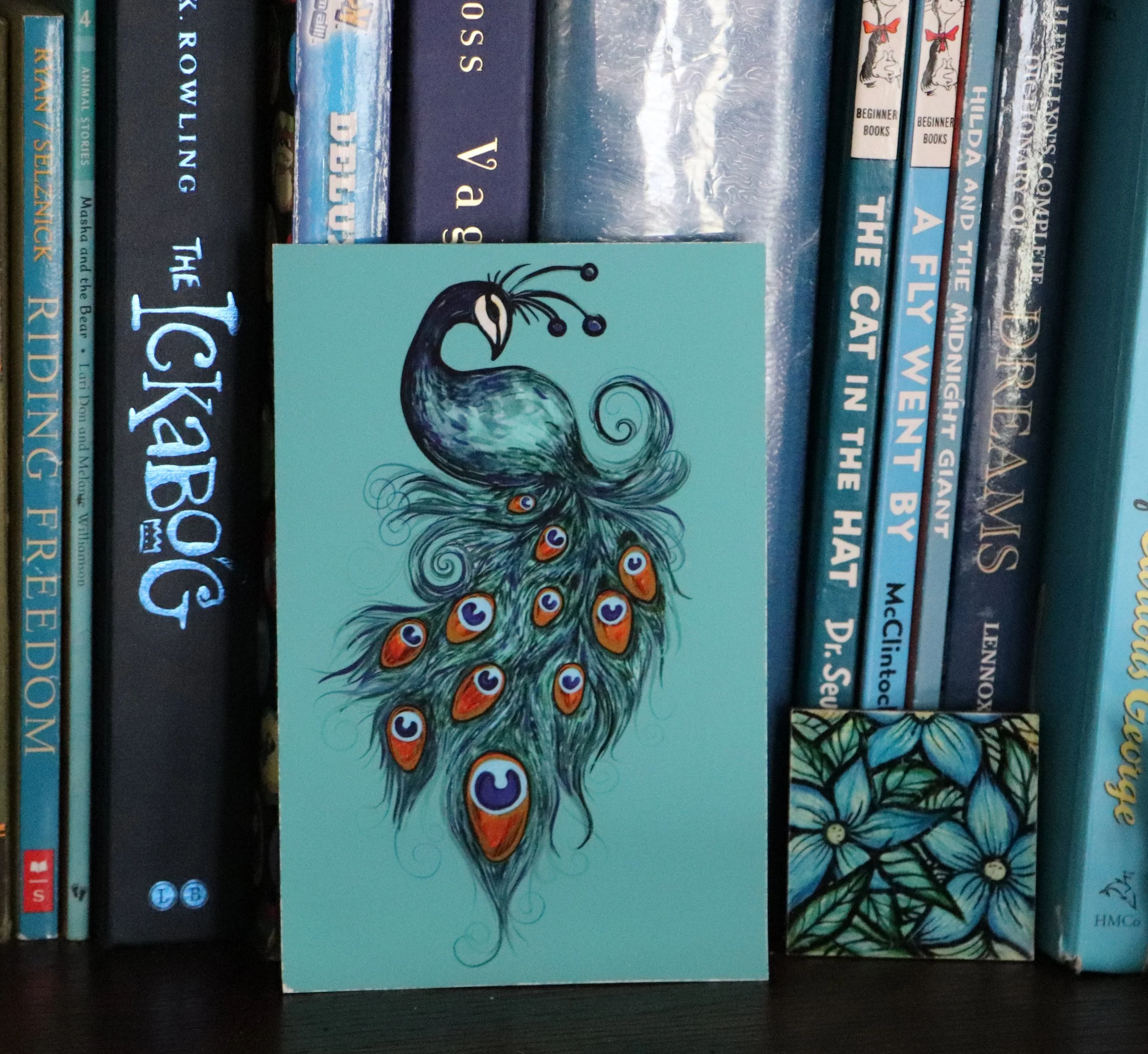 A display of books on a shelf with a peacock artwork card in front. The books are mostly blue, with titles related to animals, flying, and nature.