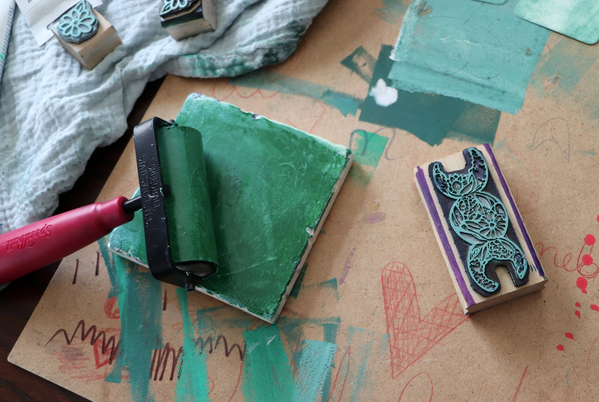 A rubber stamp with floral design, an ink roller with green ink, and a piece of paper with colorful scribbles and heart drawings on a table.