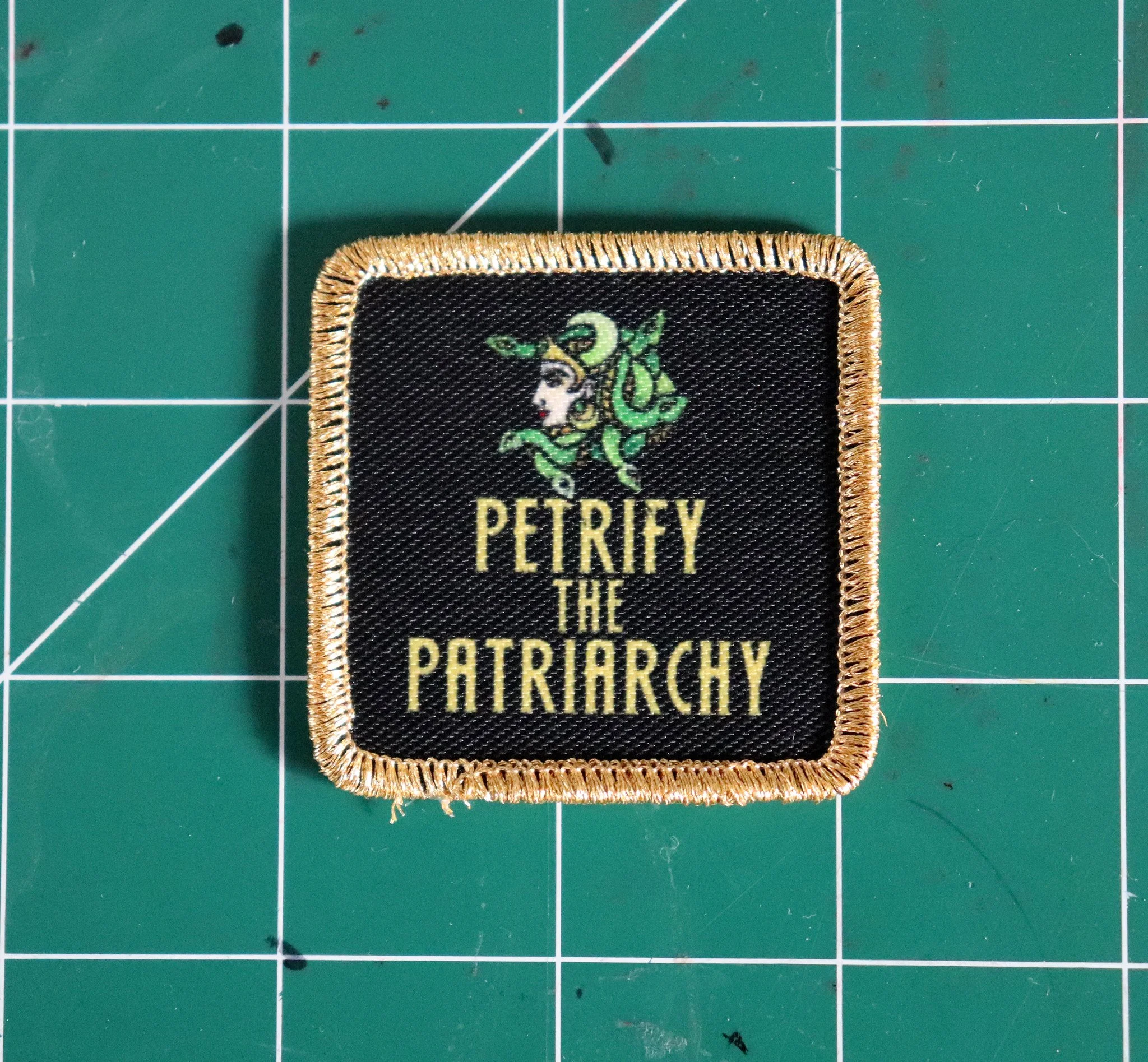 Close-up of a black embroidered patch with gold border featuring a stylized female profile with green hair and a crown, and text reading "PETROLEY THE PATRIARCHY". The patch is on a green cutting mat with white grid lines.