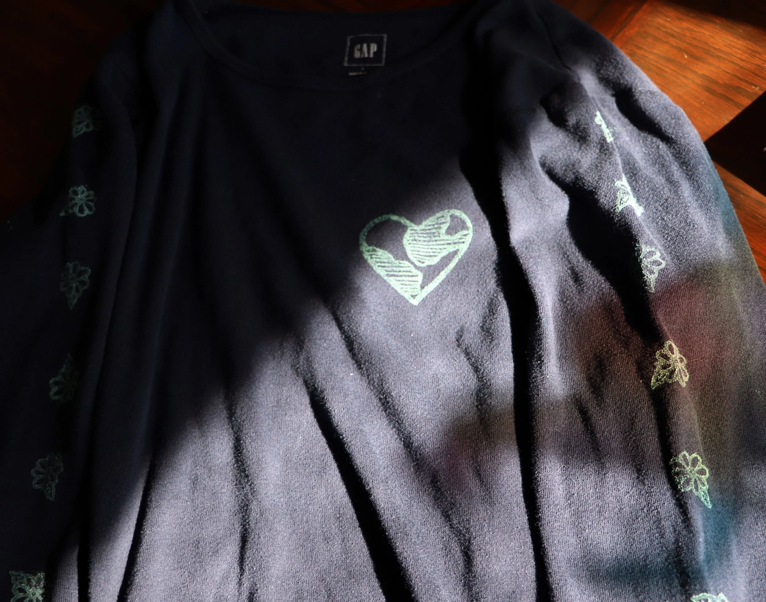 Black GAP sweatshirt with green embroidered heart and floral patterns, spread across the shoulders and sleeves, laid on a wooden surface.