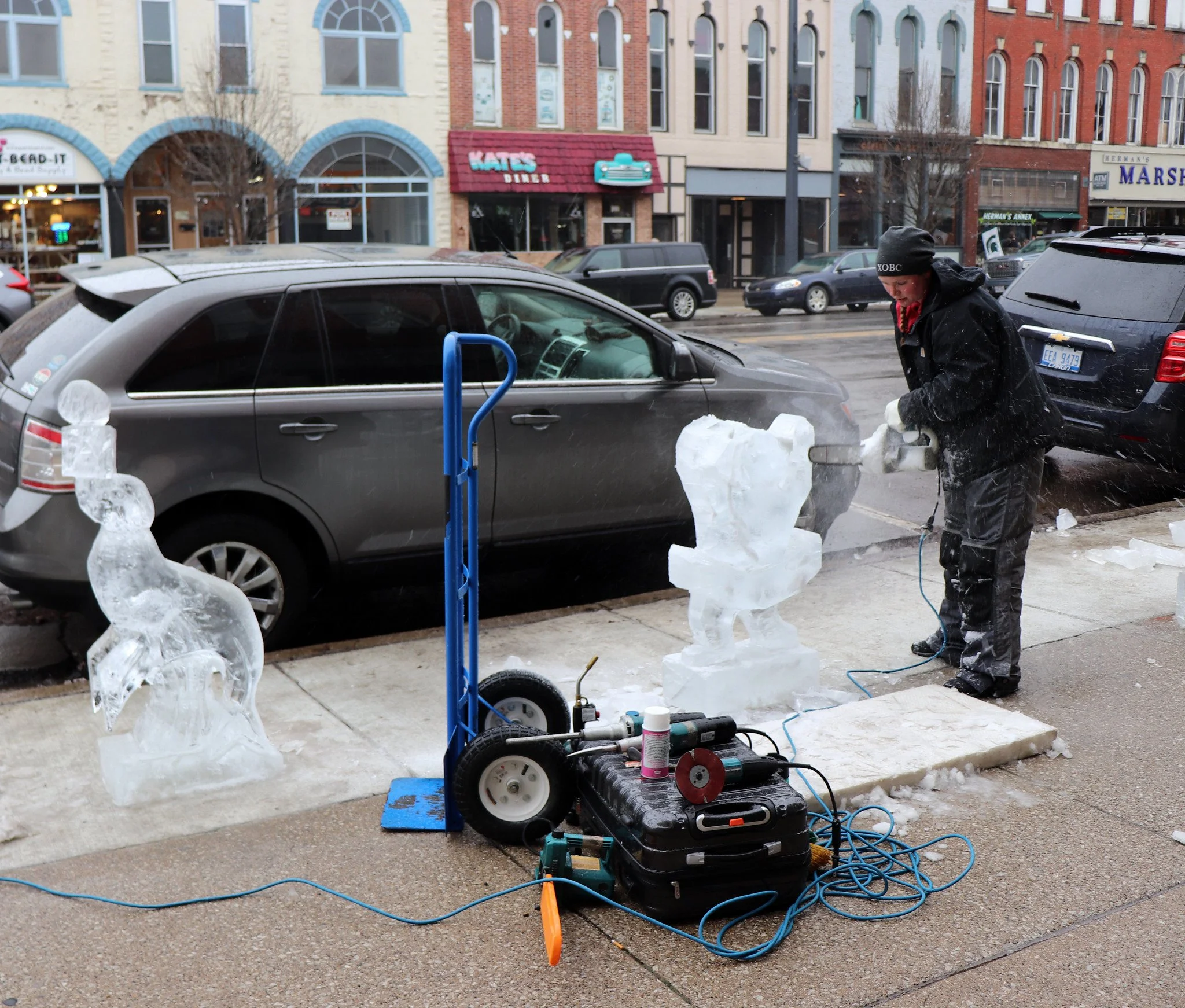 An ice sculptor carving a sculpture on a city sidewalk using a power tool, with ice figures and ice blocks around, parked cars, and storefronts in the background.