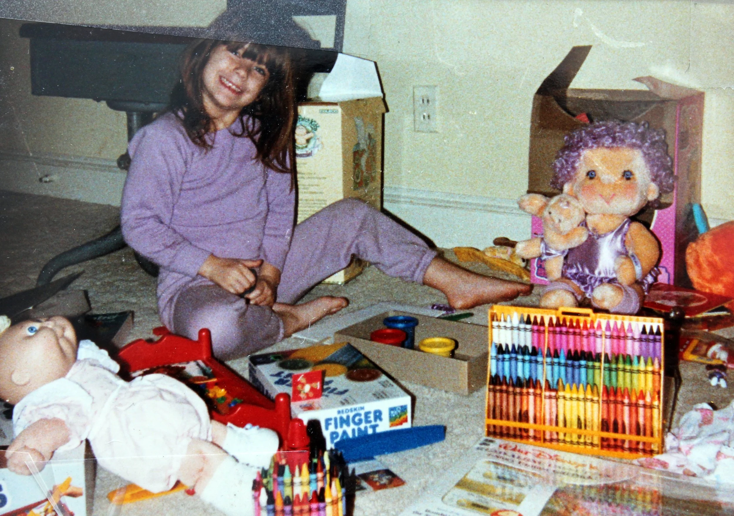 A young girl in purple pajamas sitting on the floor next to a large doll, surrounded by various toys, including colored pencils, drawing supplies, and a finger paint kit.