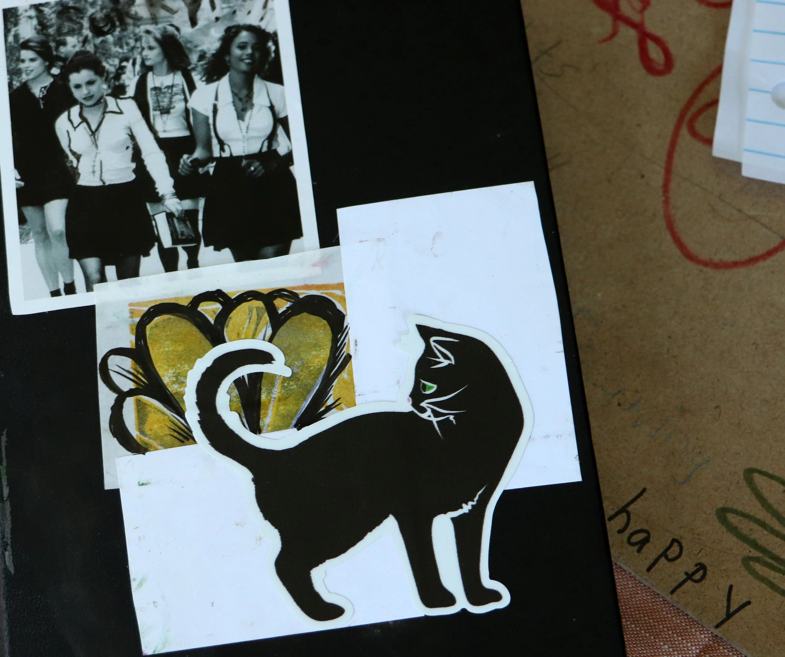 A black and white photo of five women in matching outfits, a sticker of a black cat with green eyes in front, and a colorful abstract drawing behind the cat sticker on a dark surface. Part of a cardboard surface with the words 'happy' and '...' is vi