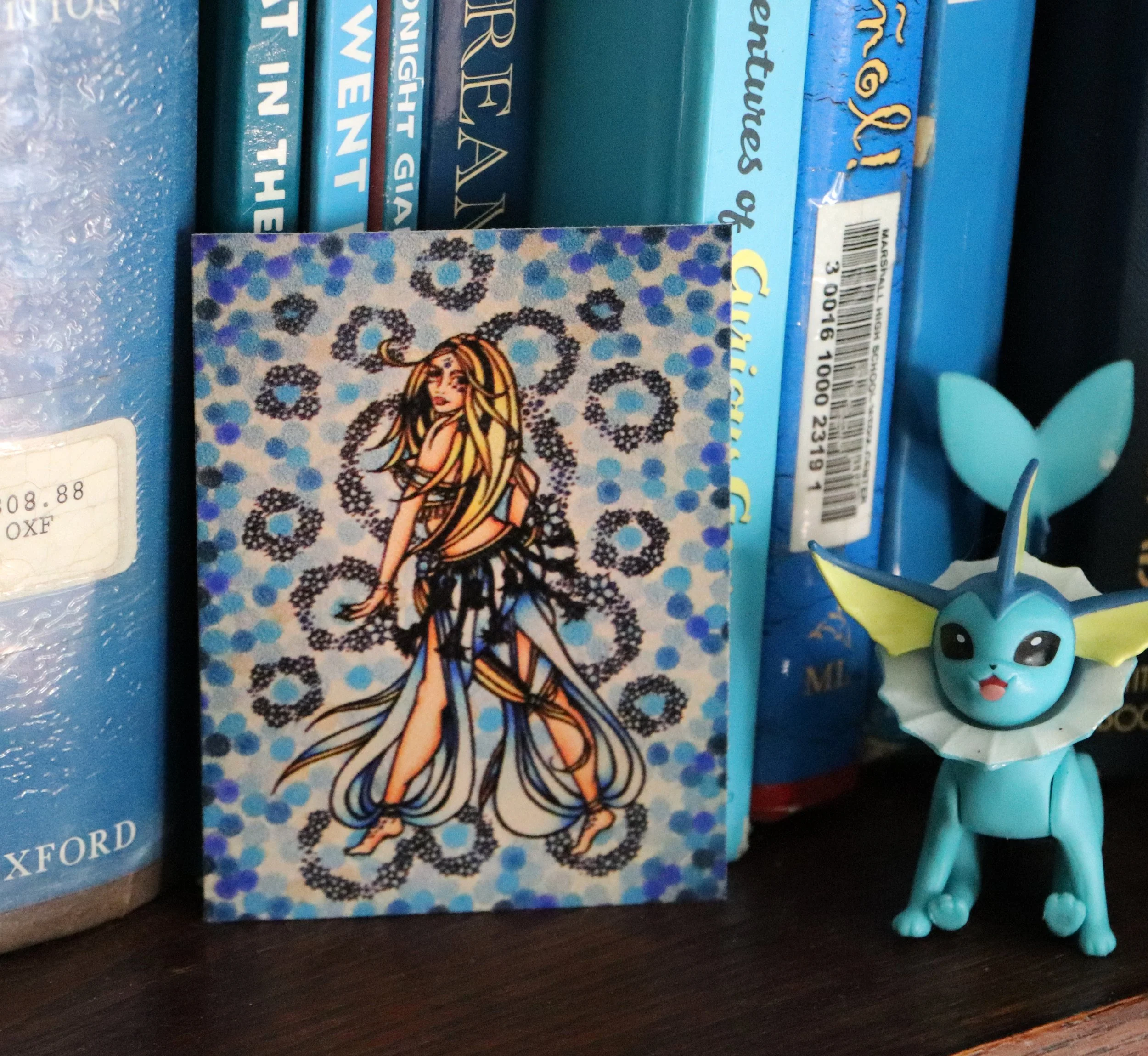 A colorful card featuring a woman with long hair, dressed in a flowing outfit, standing against a background with blue and black decorative patterns, positioned on a bookshelf next to a blue toy creature with large ears and black eyes.