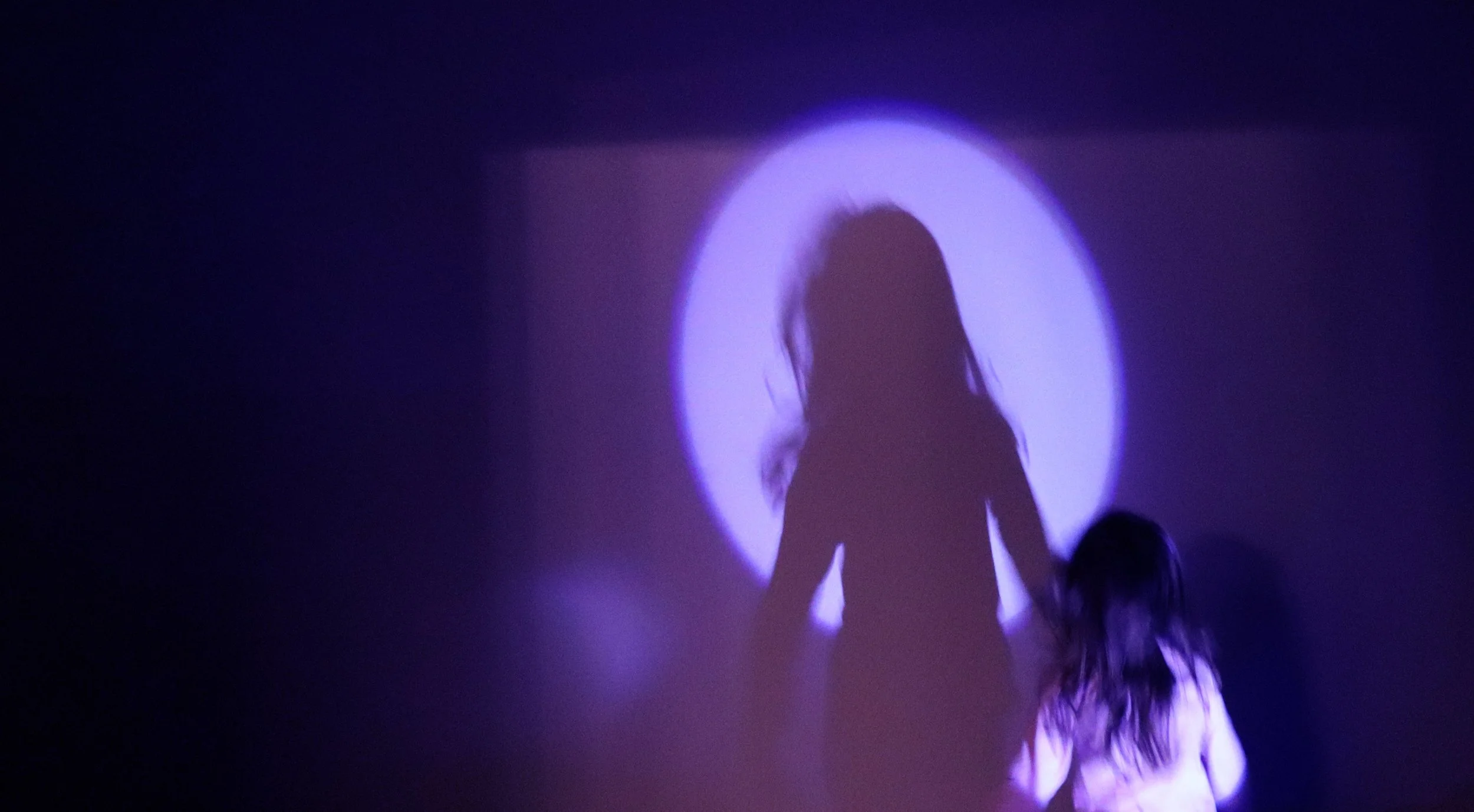 Silhouette of a young girl with long hair standing in front of a purple-lit circular backdrop.