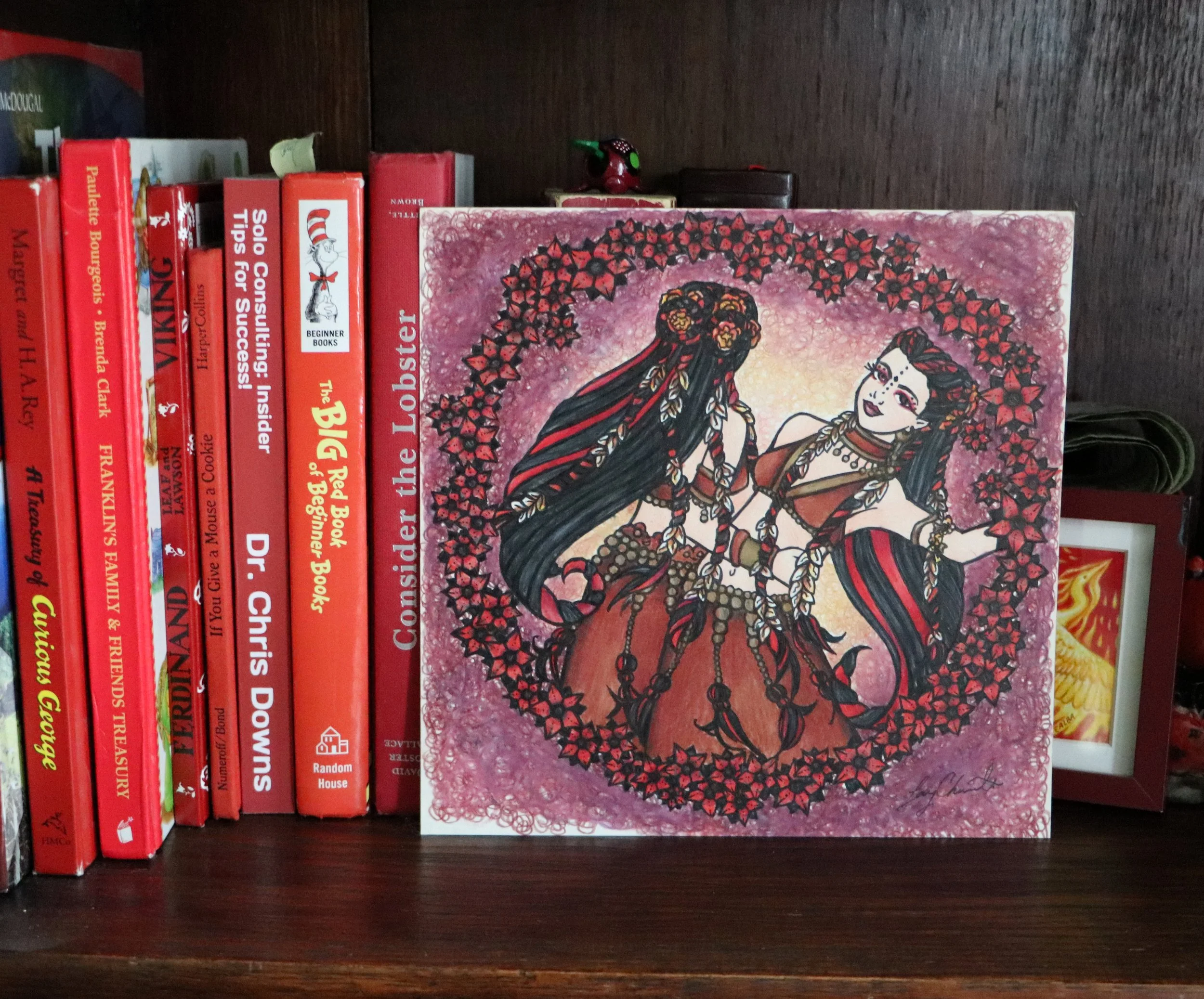 Colorful illustration of two women in traditional dresses surrounded by a floral heart shape, displayed on a bookshelf with books about writing, parenting, and books for beginners.