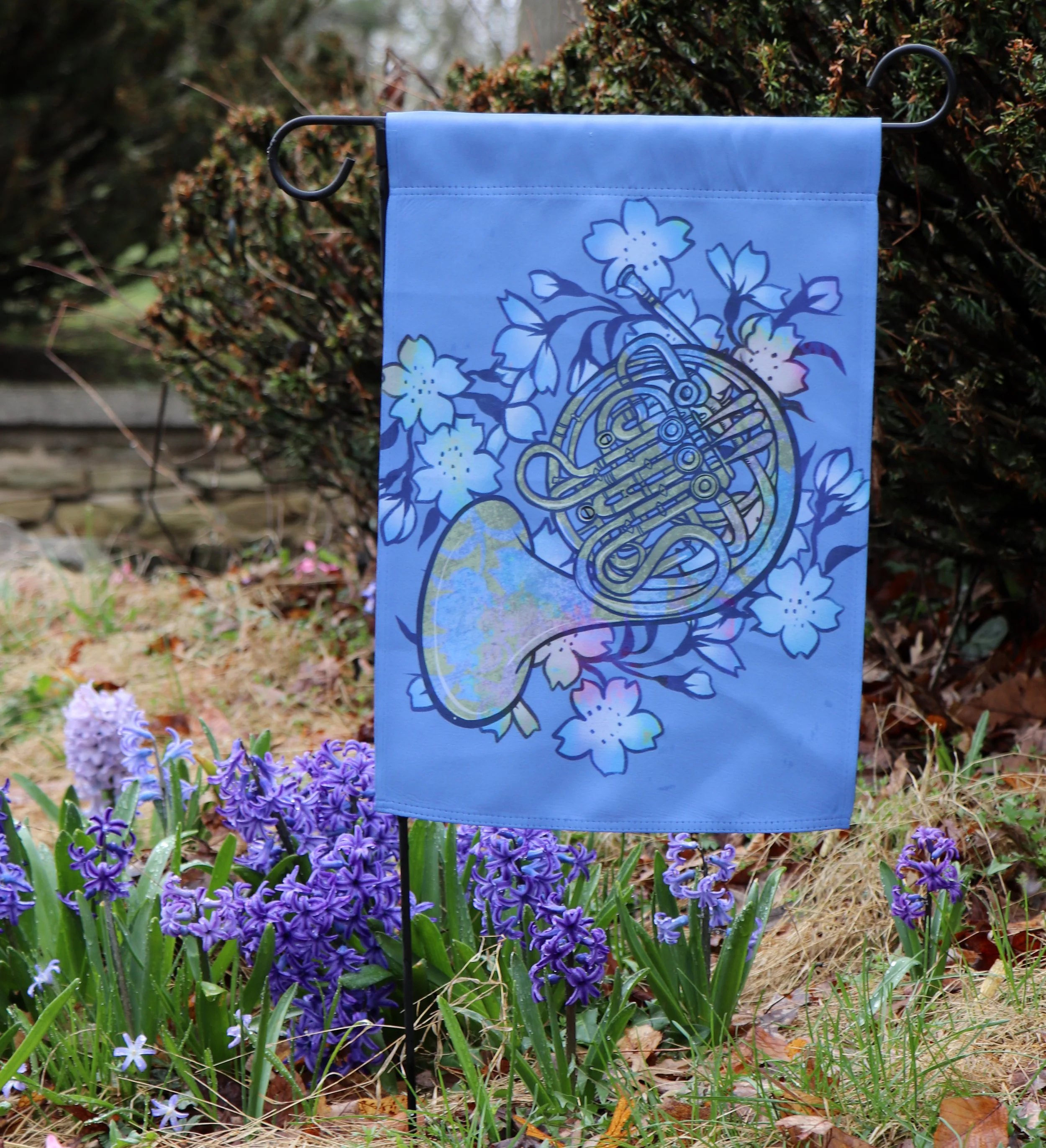 A garden flag with a floral design leaning against a bush in a garden with purple flowers and green plants.
