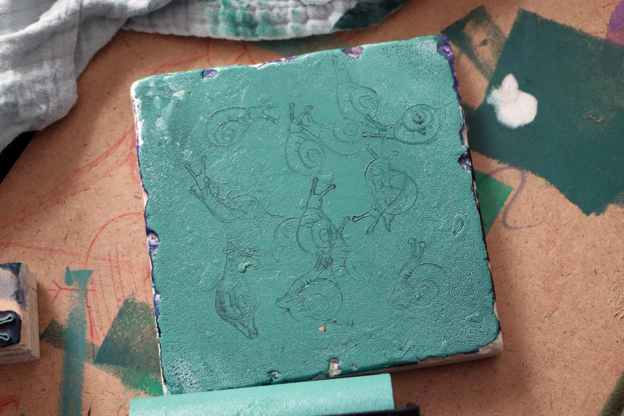 A square tile with intricate chameleon outlines engraved on its surface, placed on a cluttered workspace with fabric and paper.