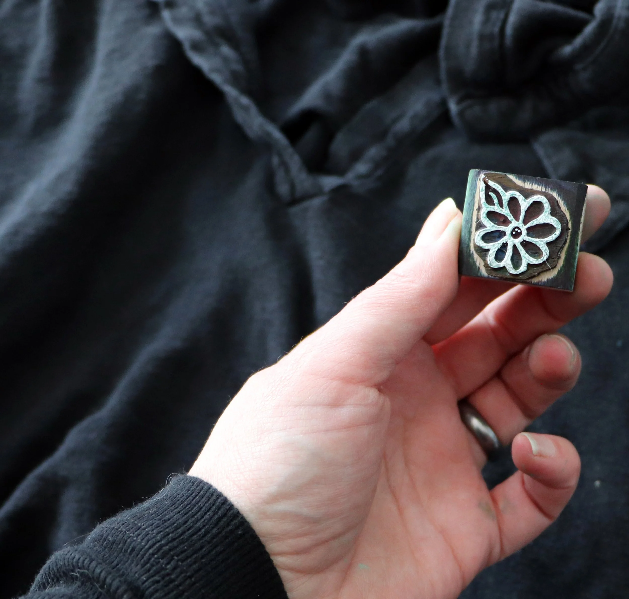 A hand holding a rubber stamp with a flower design, against a dark fabric background.