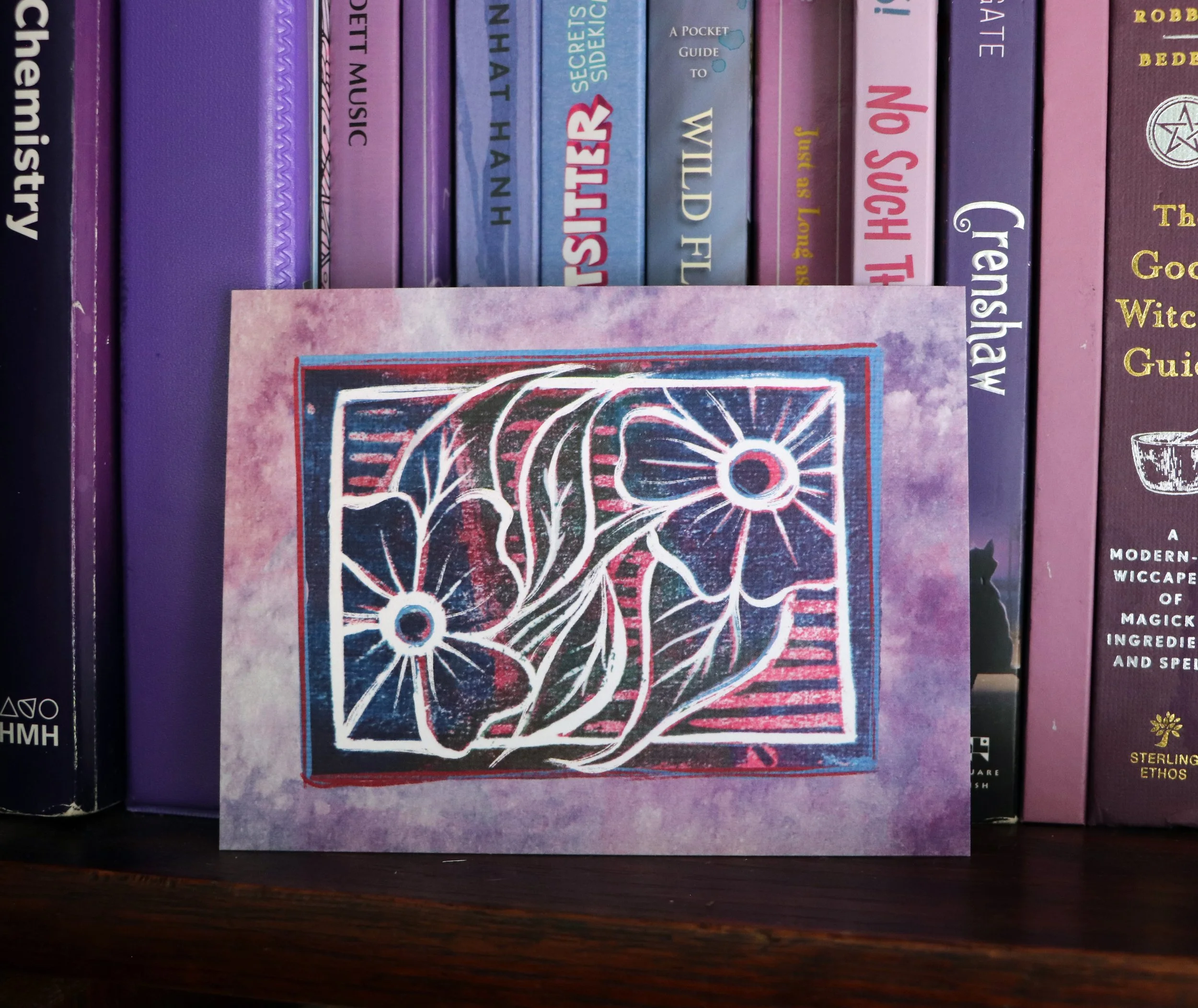A collection of books on a bookshelf, primarily purple with some pink and blue, including titles like "A Pocket Guide to" and "Witch Ethos," with a partly visible artwork or card in the foreground.