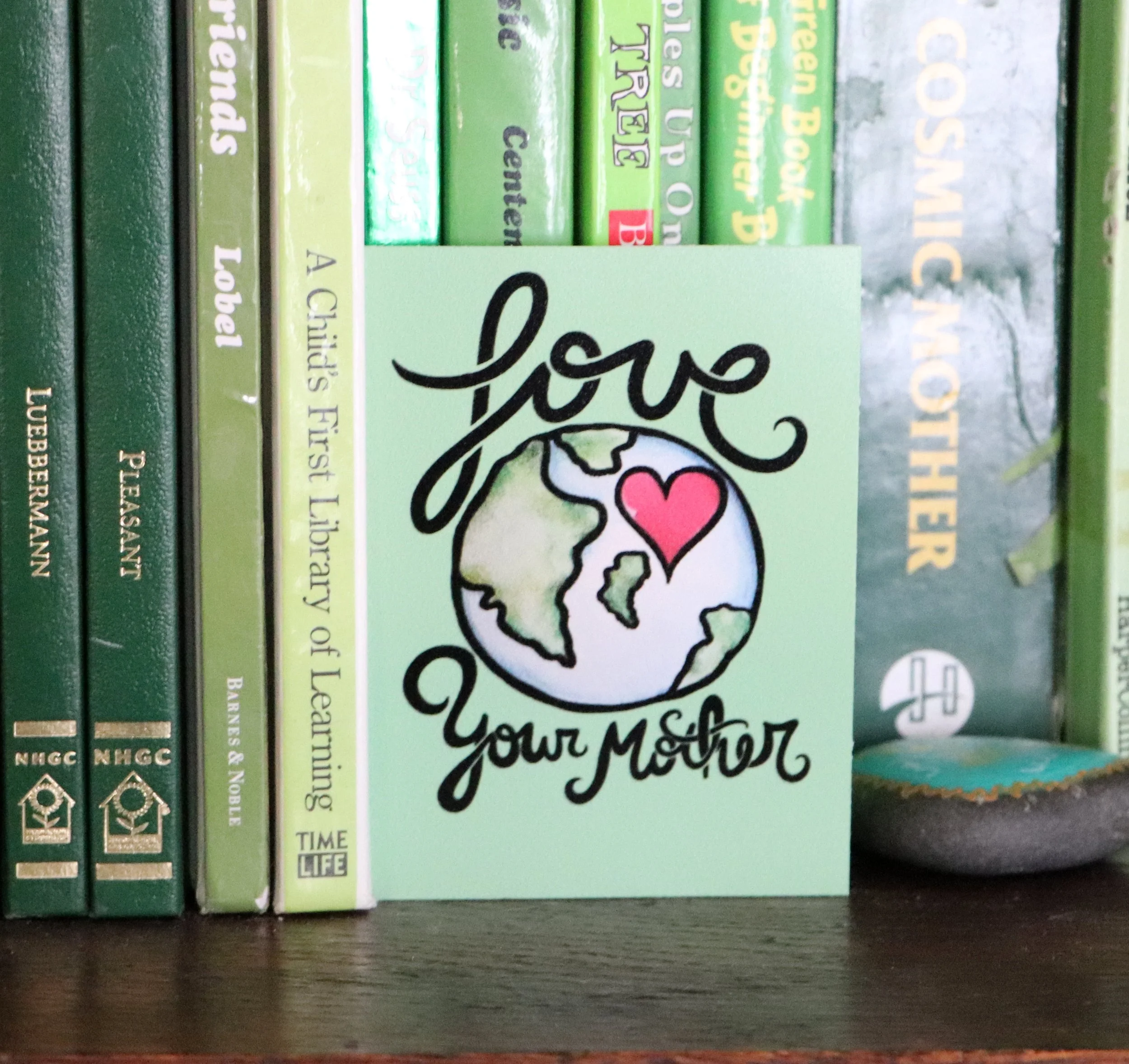 A light green card with the words 'love your mother' and an illustration of Earth with a pink heart. The card is placed on a shelf among green books.