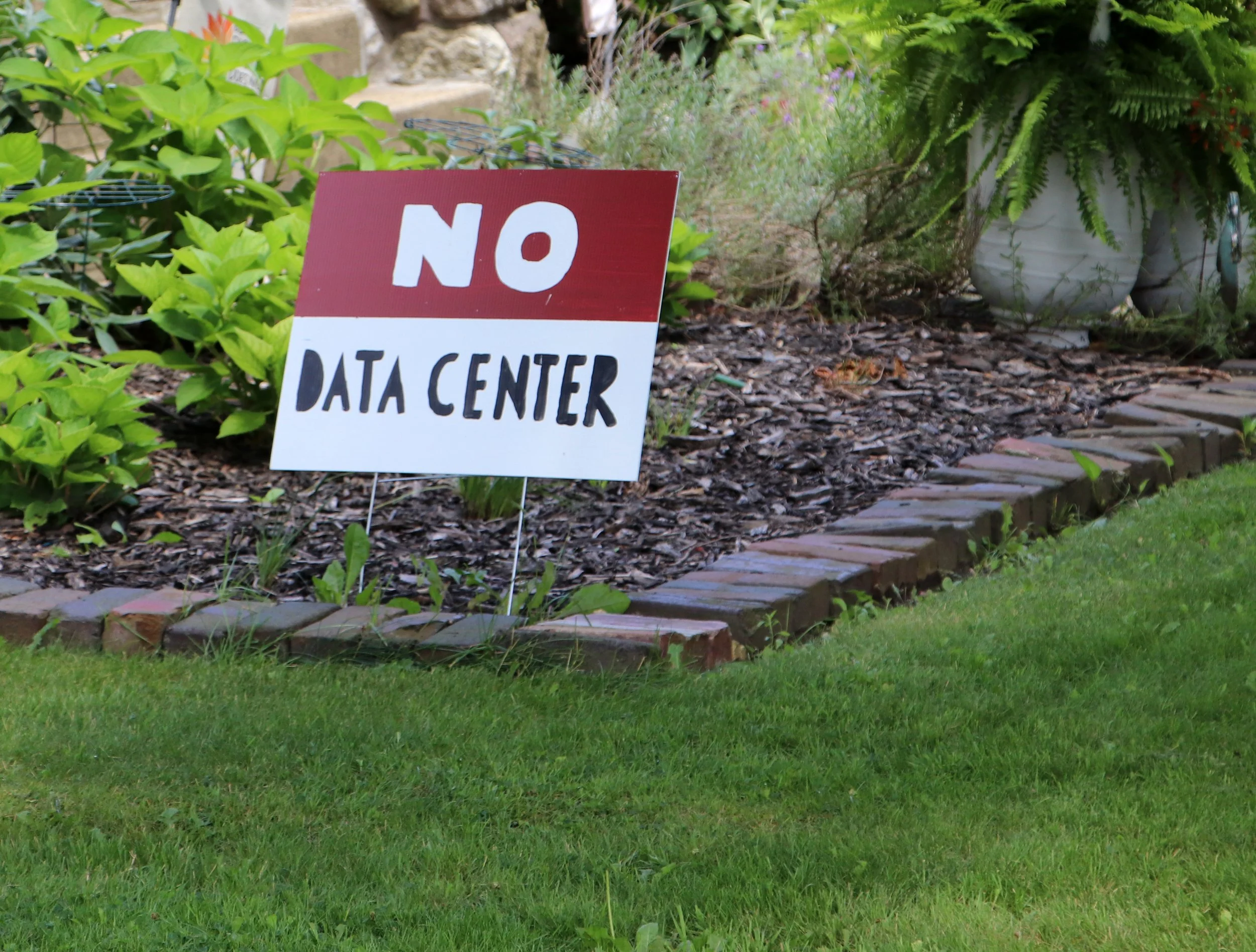 Garden with a red and white sign that reads 'NO DATA CENTER', various plants, ferns, a brick pathway, and a white decorative pot.
