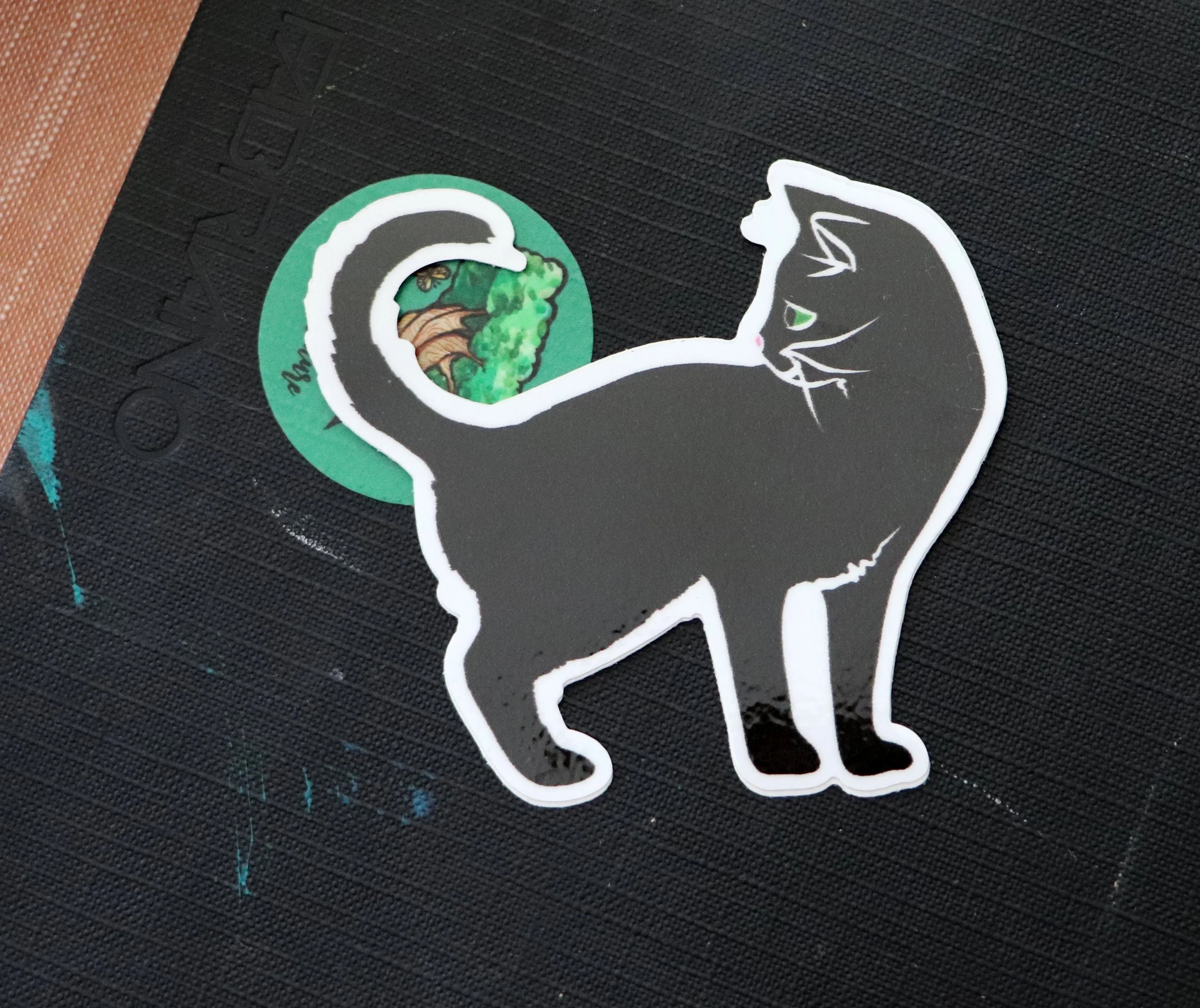 A sticker of a black cat with a white outline placed on a black textured surface with a green circular logo in the background.