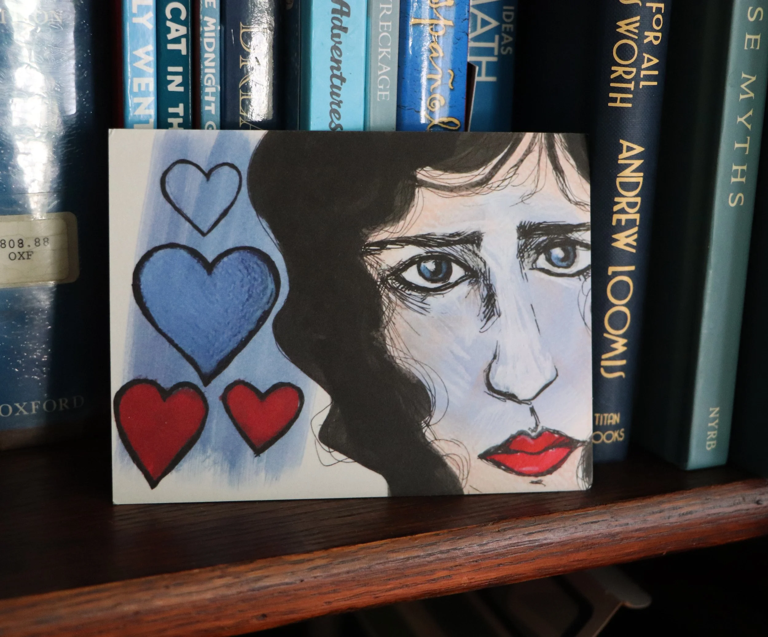 A collection of books on a shelf, featuring titles related to psychology and fiction, with a book cover showing a drawing of a woman with blue eyes, dark hair, and red lips, accompanied by hand-drawn hearts.