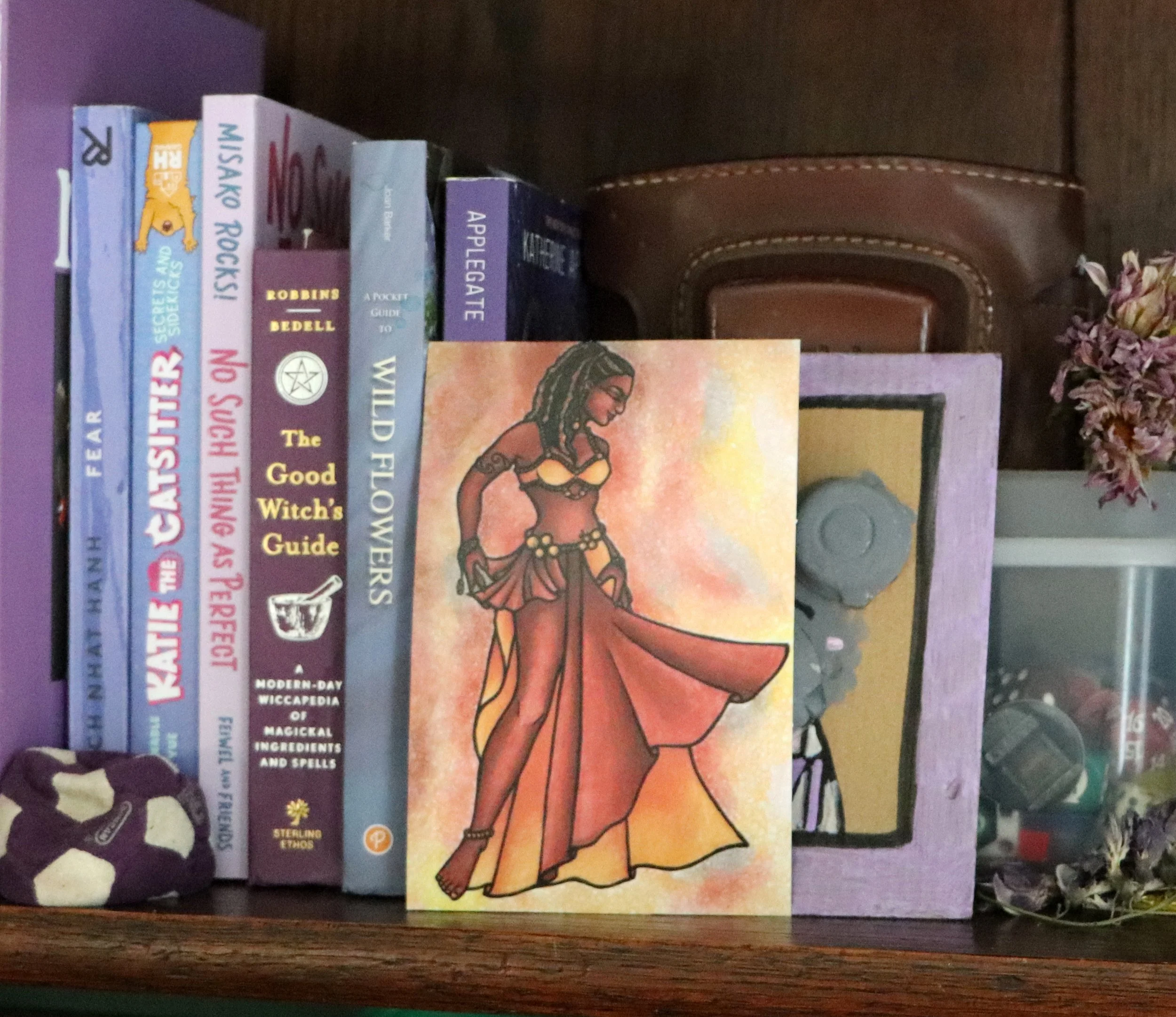 Bookshelf with fantasy, witchcraft, and nature-themed books, a framed artwork of a woman in a bohemian outfit, a purple picture frame, a small football, and a dried flower arrangement.