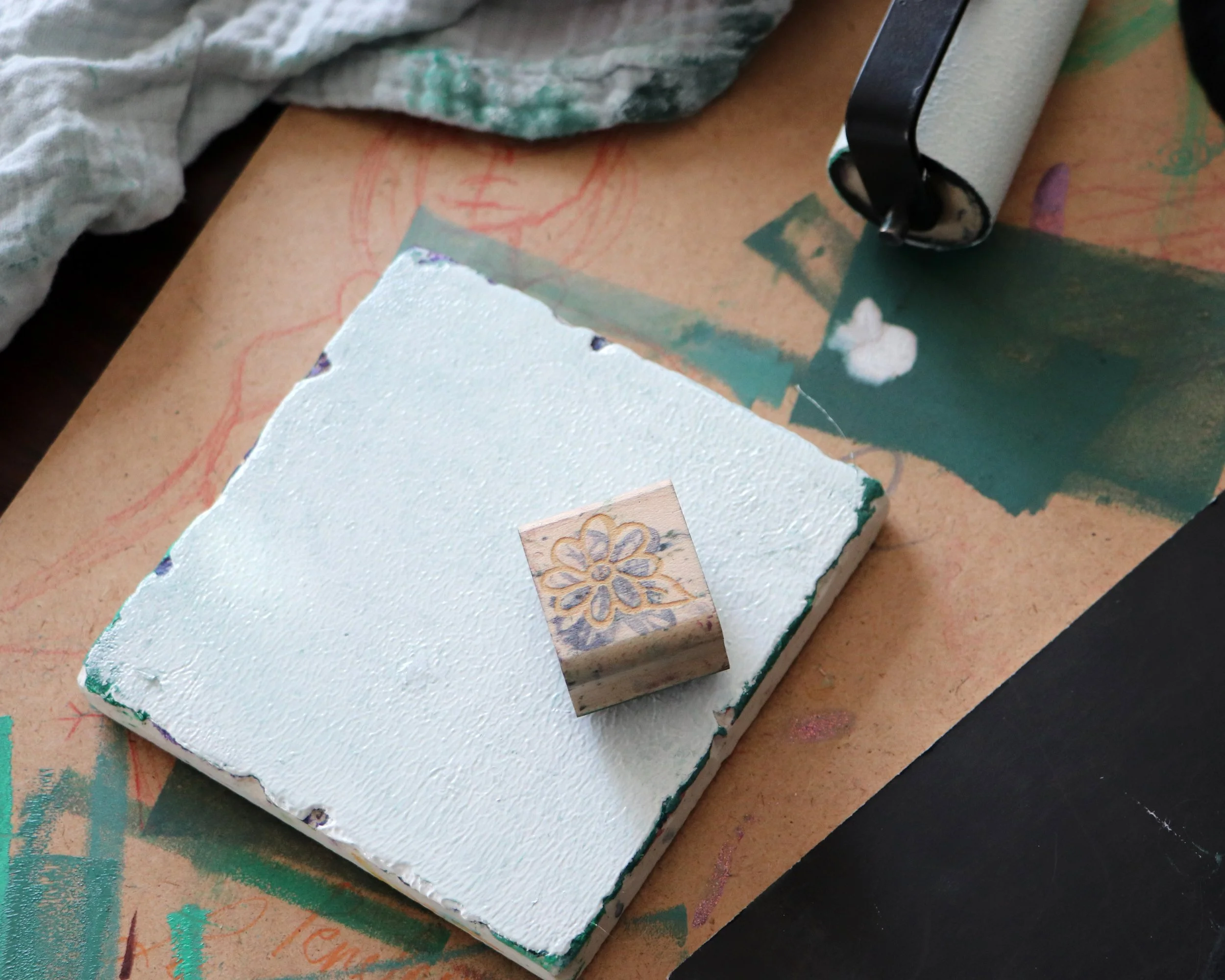 A piece of textured white foam with a small rubber stamp featuring a floral design resting on top of it, surrounded by art supplies and paper on a work desk.