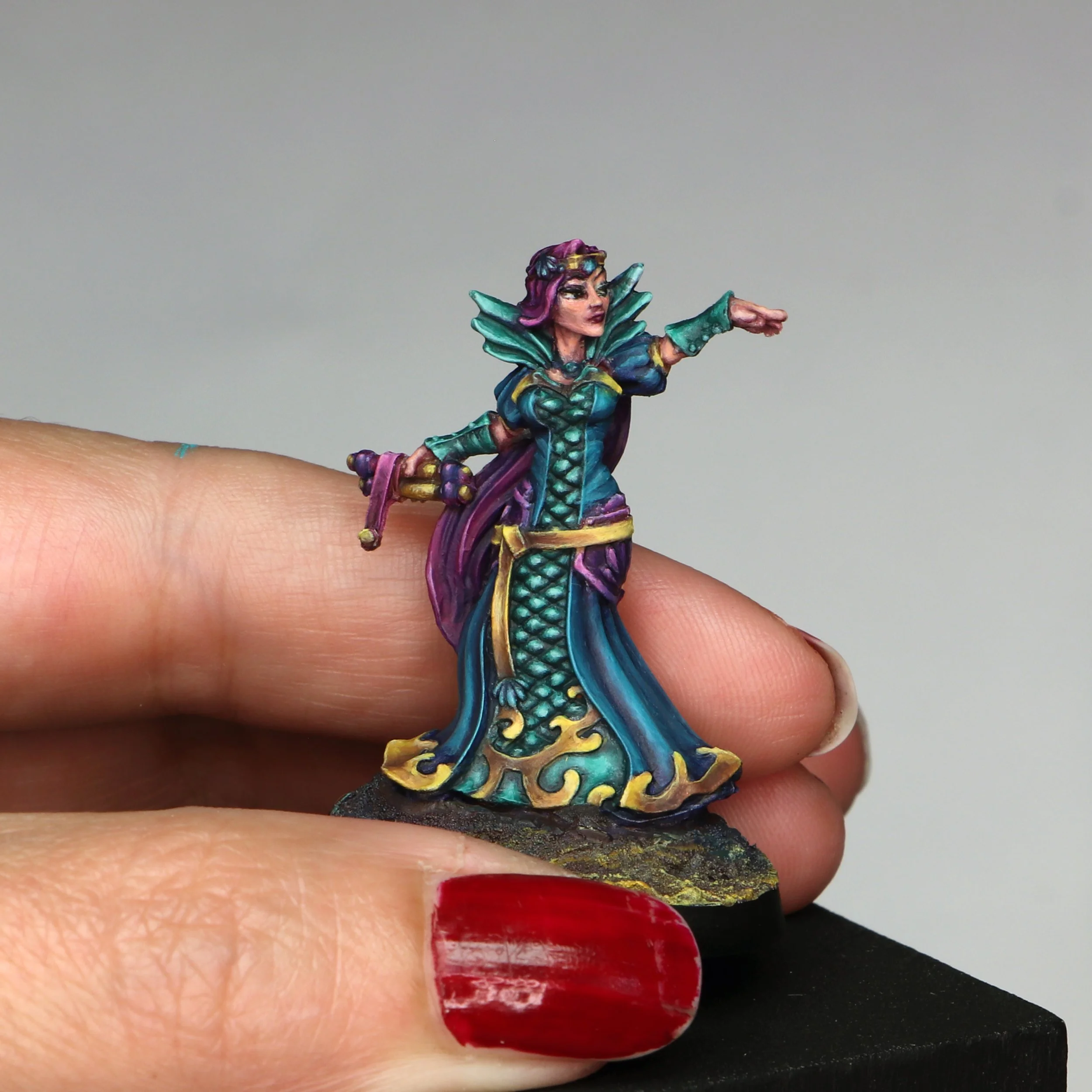 Close-up of a hand with painted nails, wearing a colorful fairy or elf miniature figure, with a plain gray background.