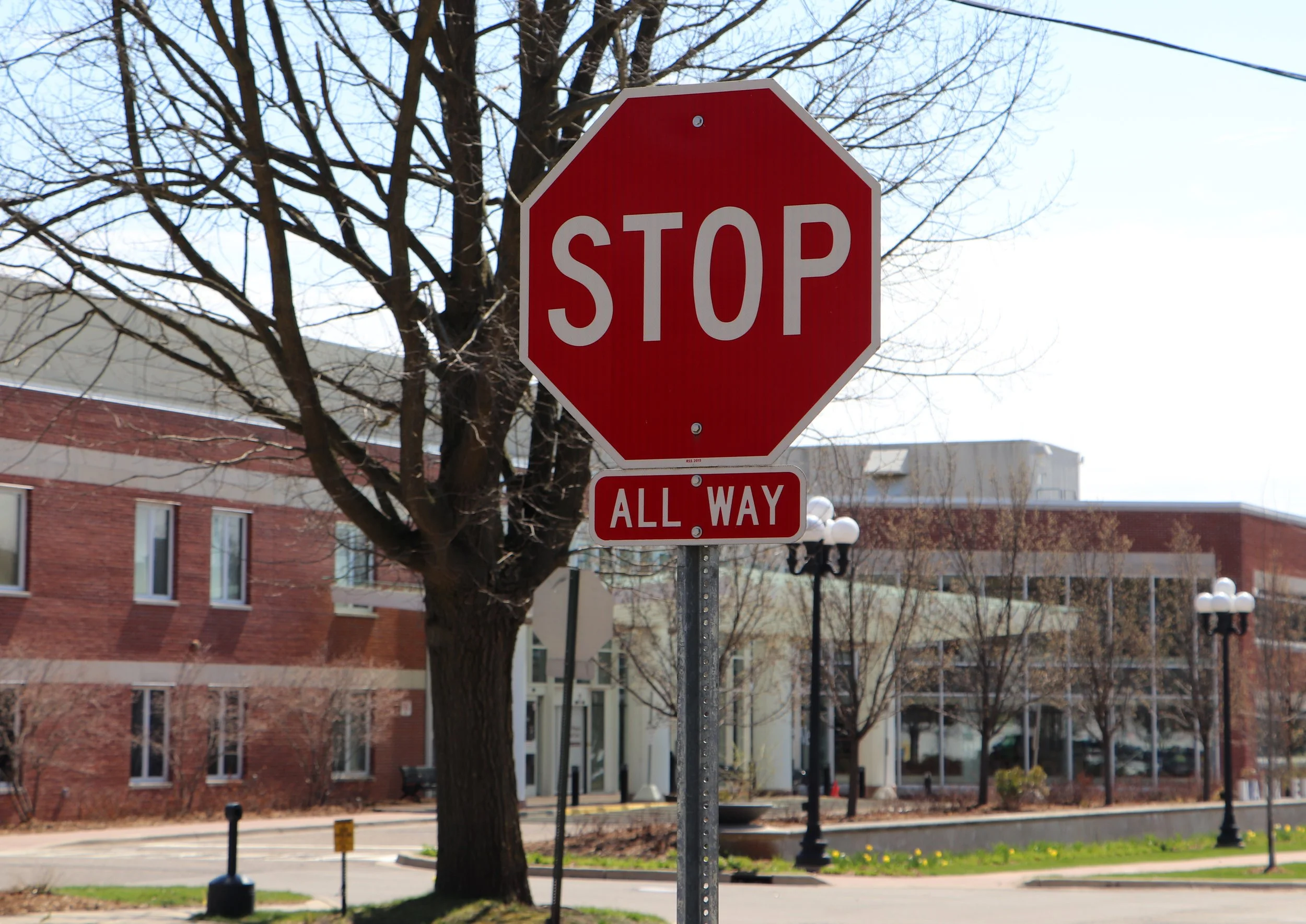 A red stop sign with white text that says STOP, positioned on a street corner. The background includes leafless trees, brick and white buildings, and a clear sky.