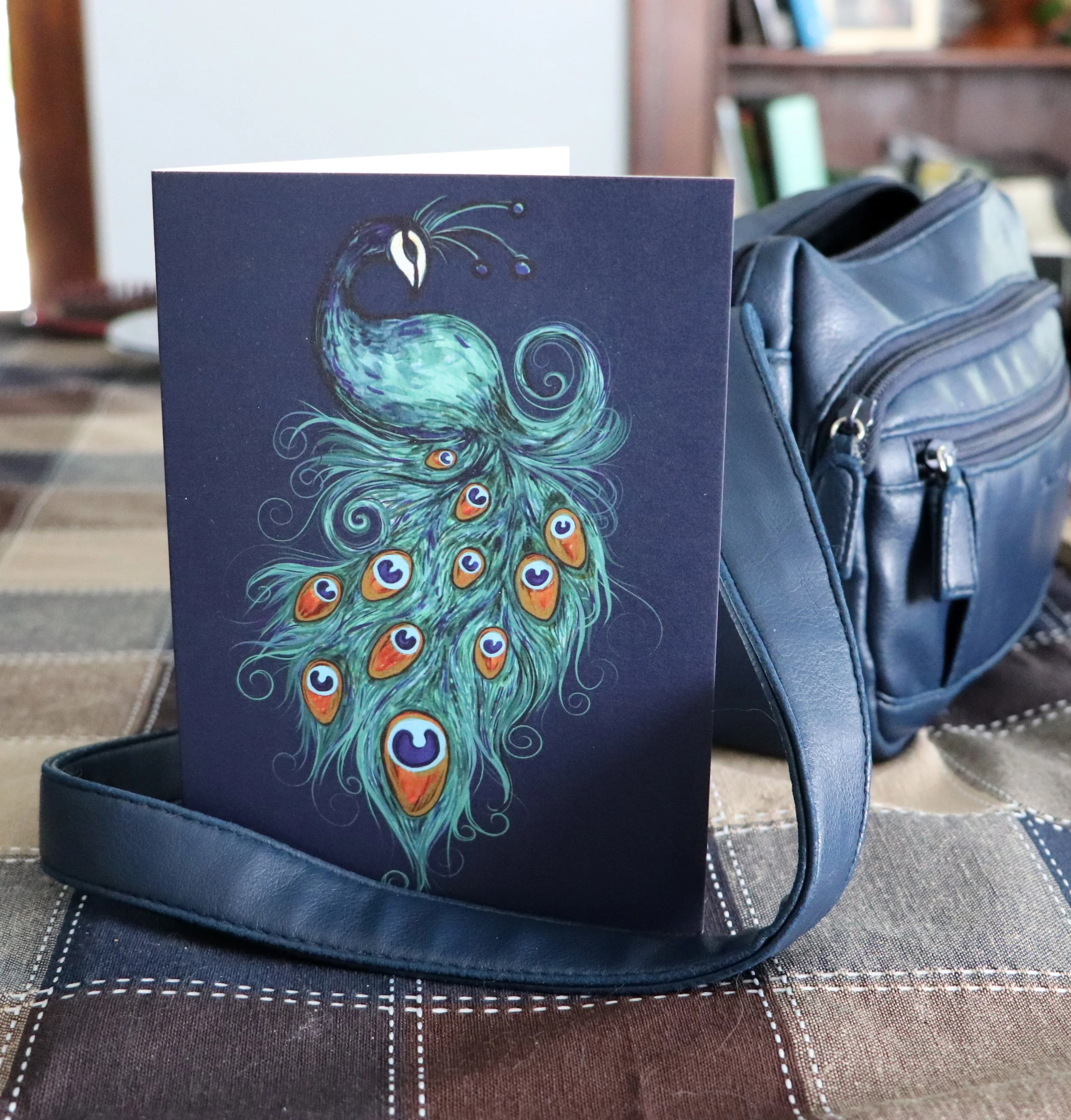 A colorful illustrated greeting card featuring a peacock with its tail fanned out, displaying blue, green, orange, and purple eye patterns, placed on a blue handbag on a patterned table cloth.