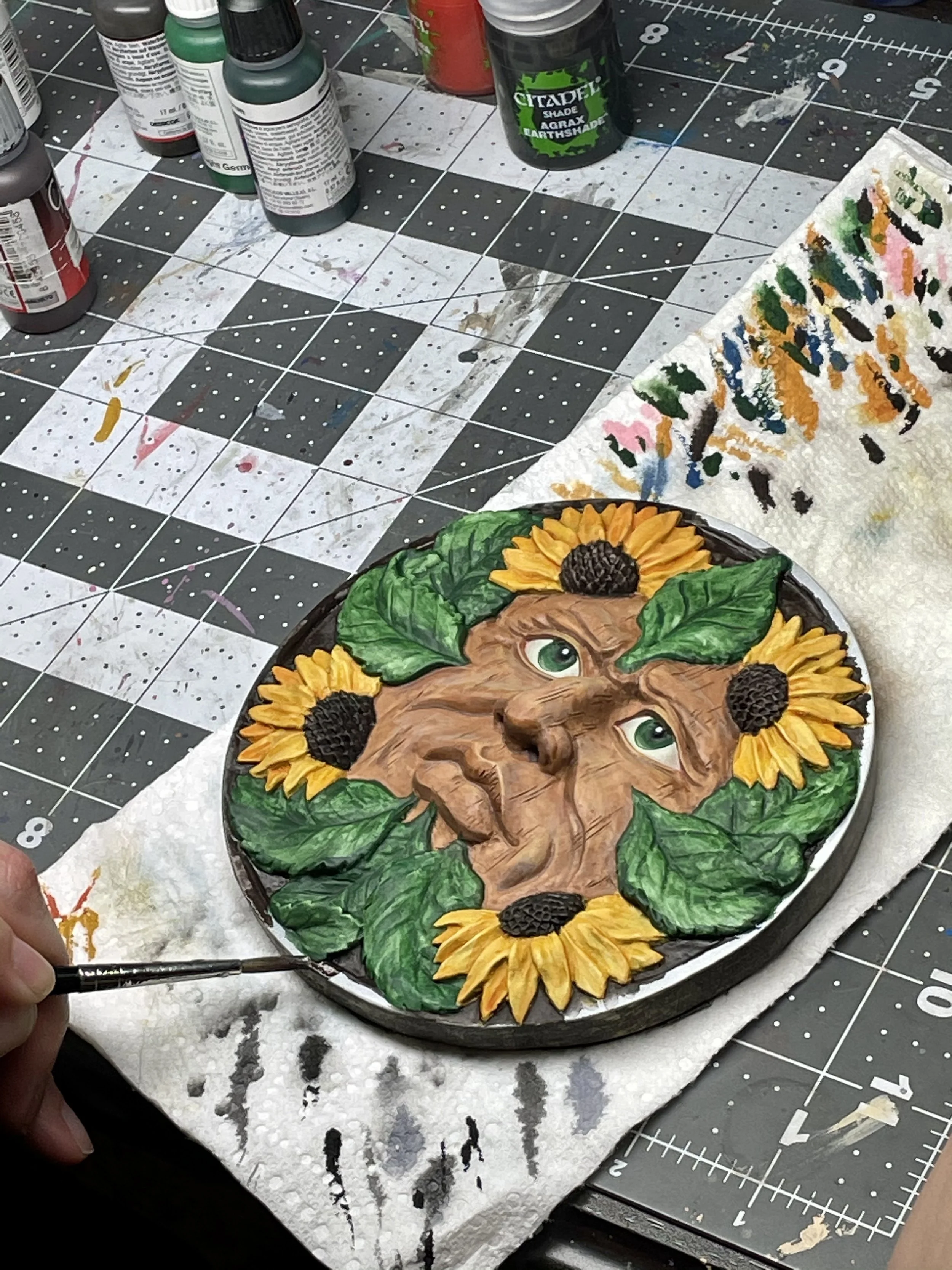A painted circular artwork depicting a face with green eyes, surrounded by yellow sunflowers and green leaves. The scene is set on a work surface with art supplies and a paper towel nearby.