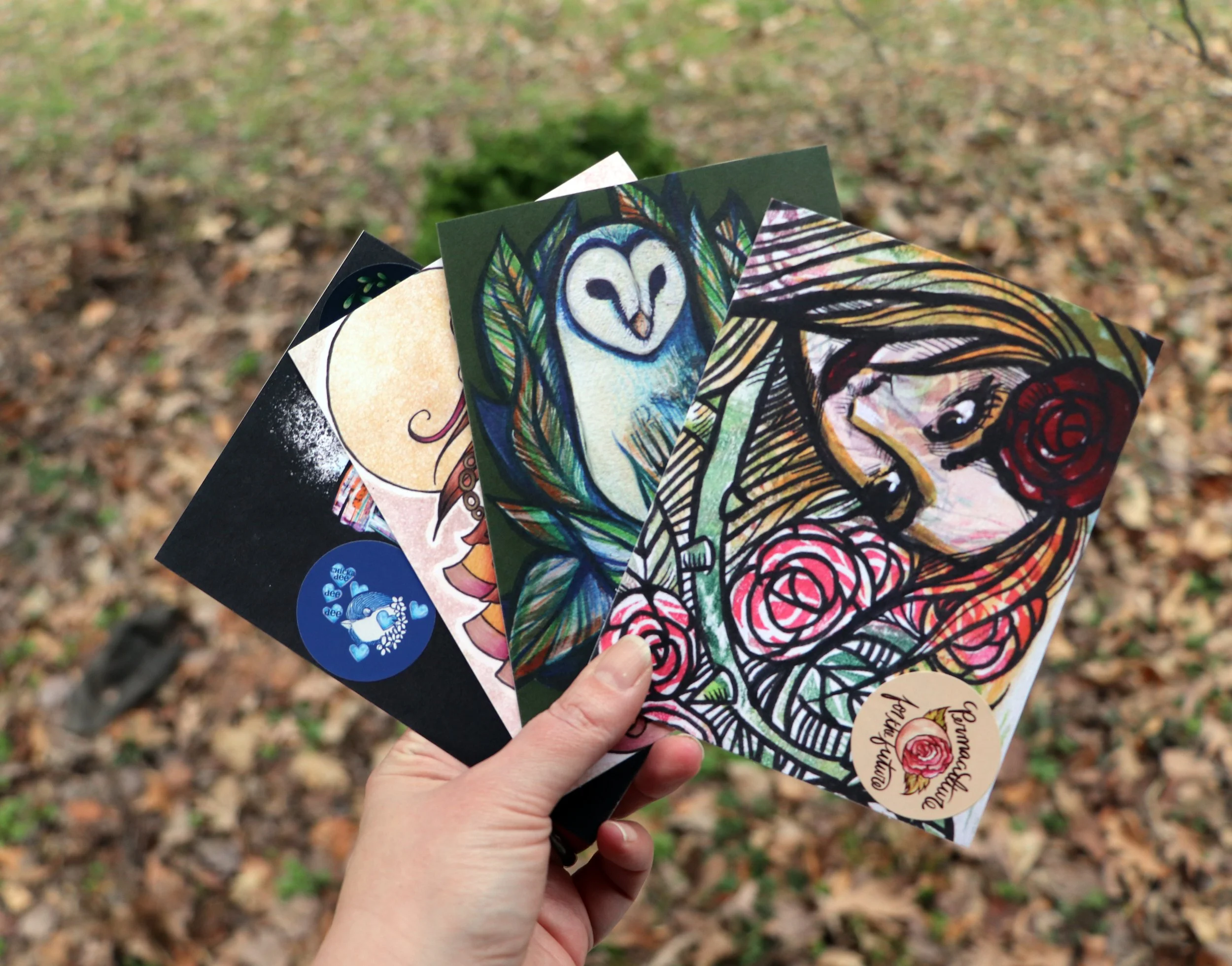 Hand holding four handmade illustrated postcards outdoors on a background of fallen leaves and grass.