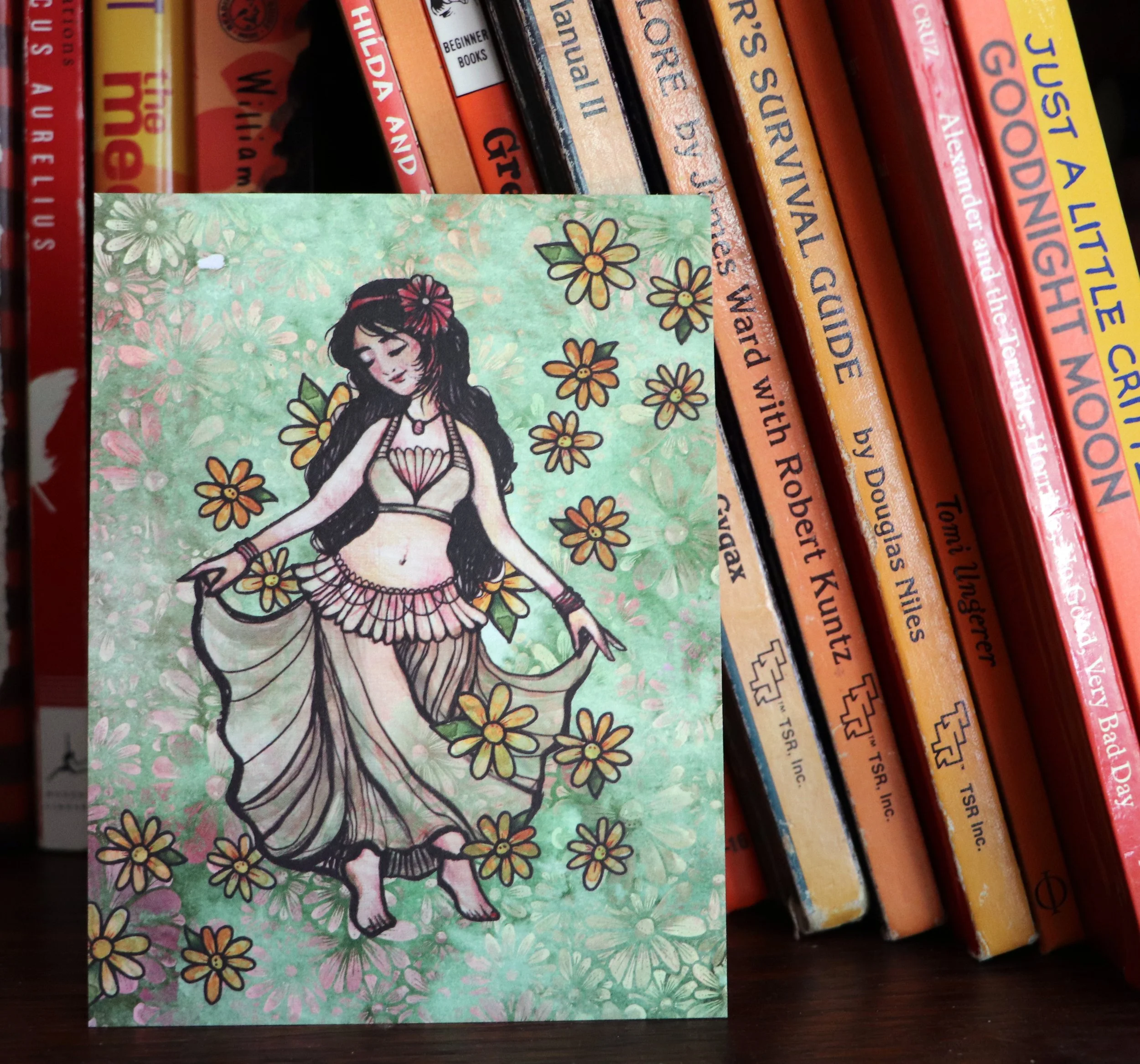 A collection of books with colorful covers and titles, a small illustrated card featuring a girl with long dark hair, surrounded by flowers, is placed in front of the books.