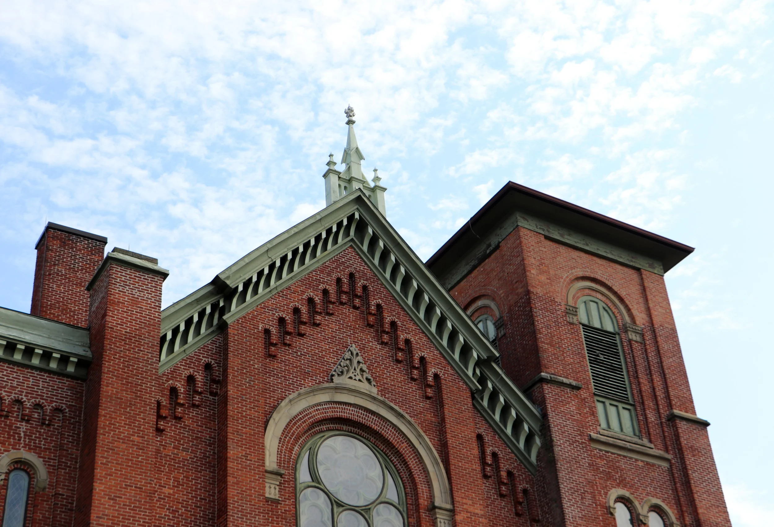 The top section of a red brick church with a pointed steeple and ornate architectural details, set against a partly cloudy sky.