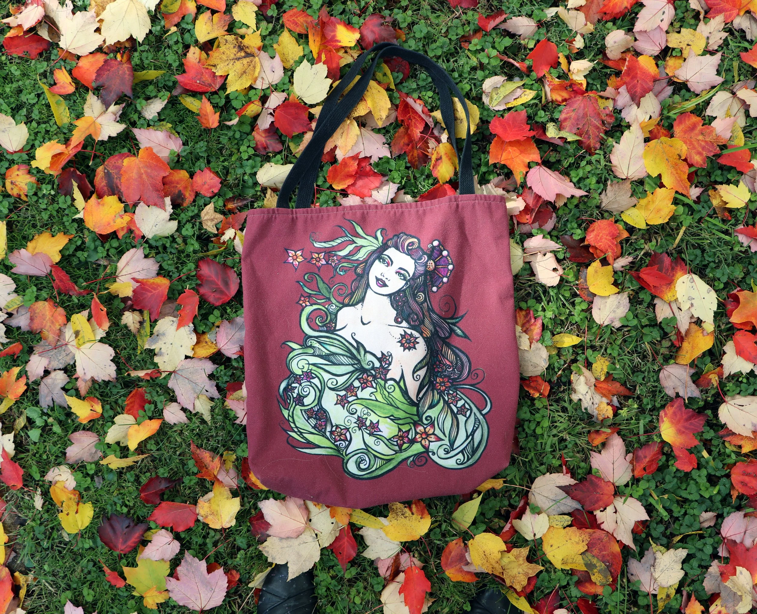 A maroon tote bag with a colorful illustration of a woman with long flowing hair, surrounded by floral and leaf designs, lies on the ground among fallen autumn leaves in shades of red, yellow, and orange. Green grass and small plants are visible bene