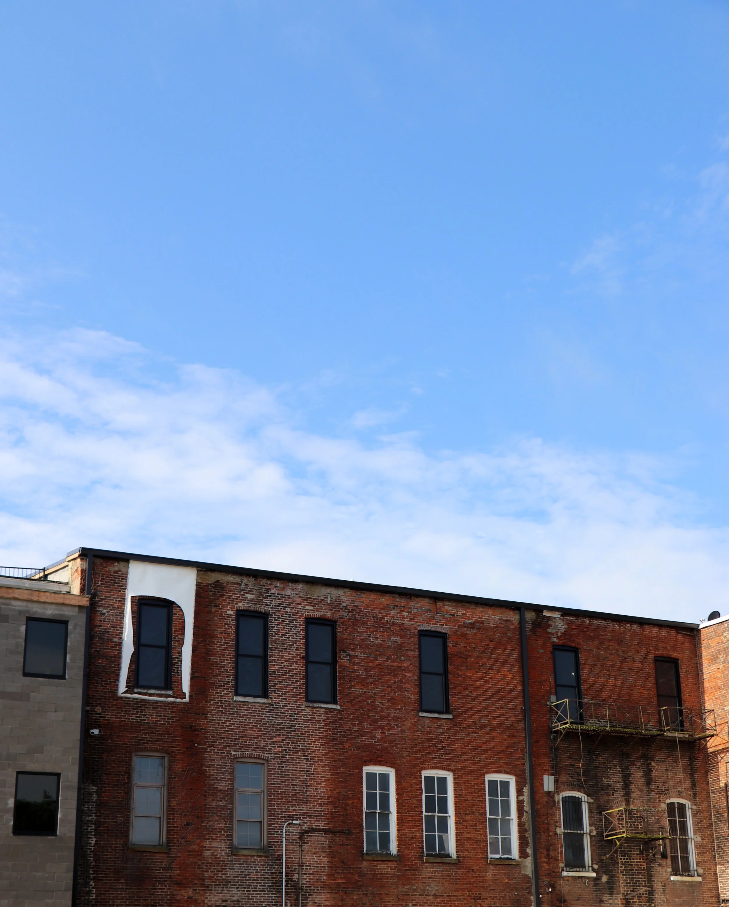 A city building with multiple windows, brick and gray facade, against a bright blue sky with a few clouds.